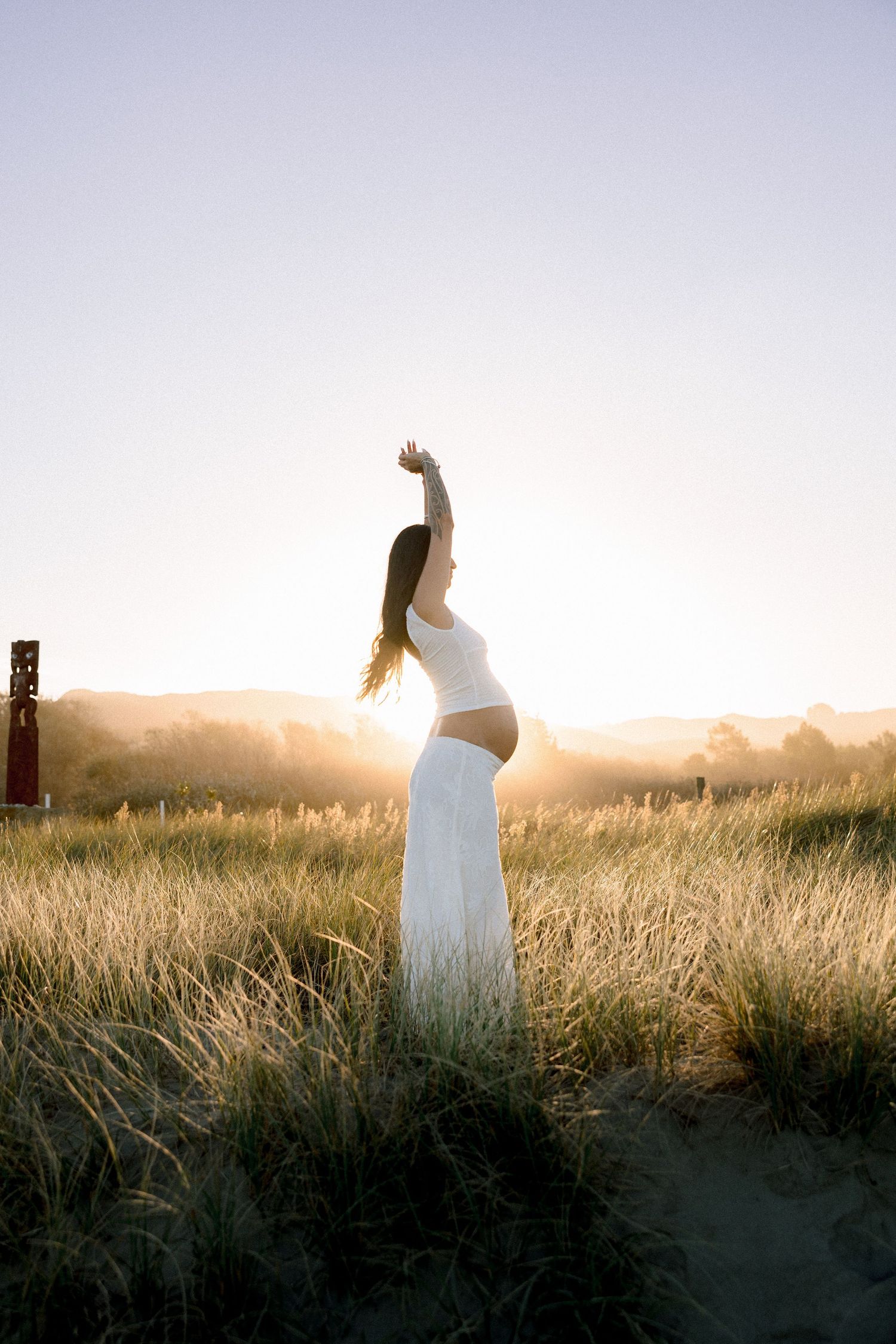 Pregnant woman in white dress stands in golden sunset field, arm raised gracefully above head.