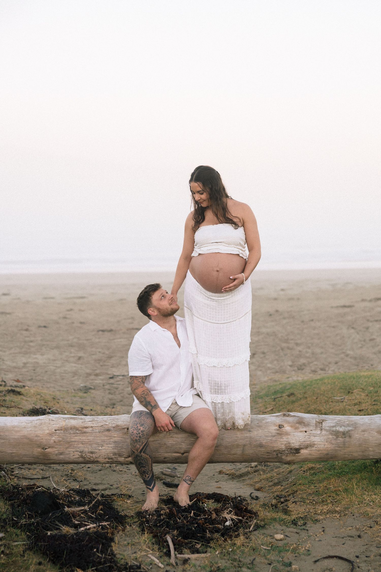 Couple at beach maternity shoot, man sitting on driftwood touching pregnant woman's belly, both wearing white.