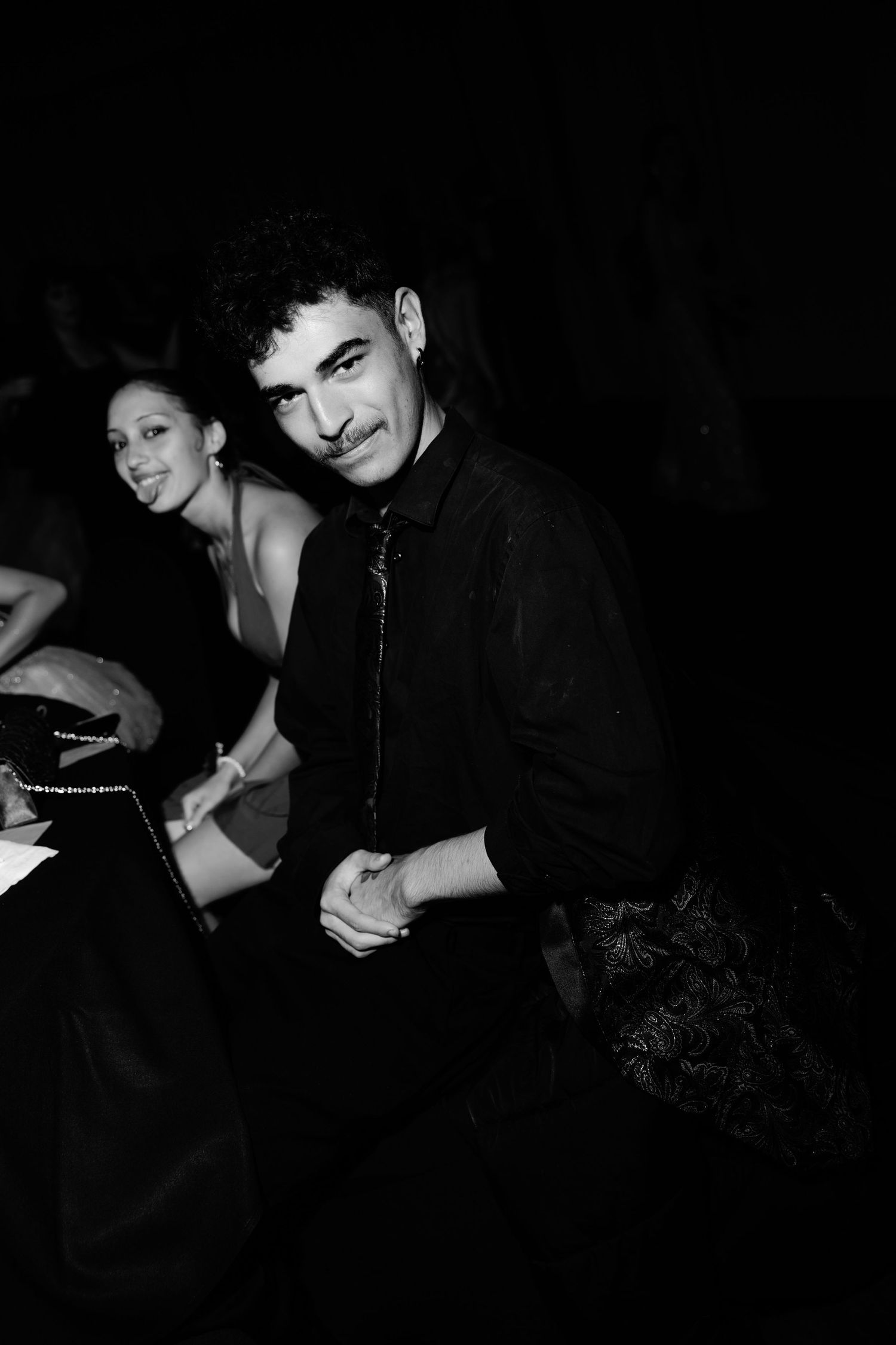 Black and white photo of a man with mustache in black shirt and tie posing with a woman at a formal event.
