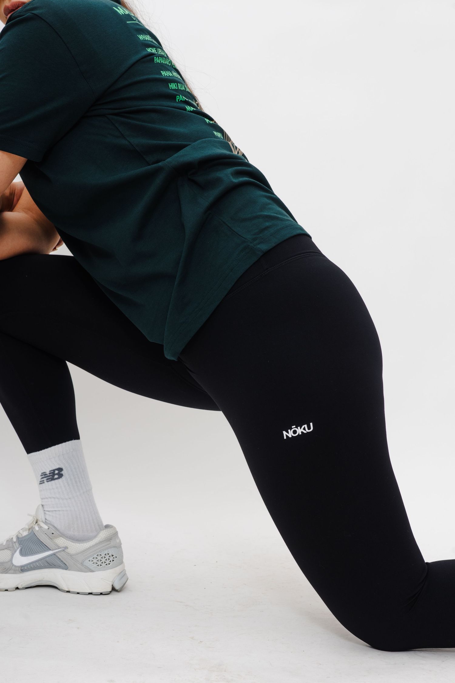 Close-up of person wearing black leggings with NOAD logo, dark green top and gray Nike sneakers in lunging position.