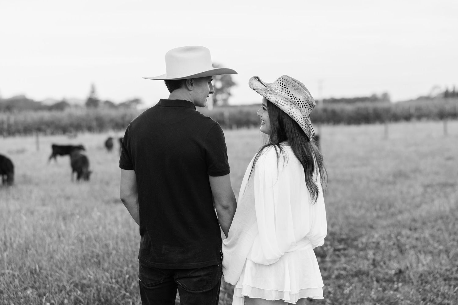 A couple in wedding attire walks hand in hand through a scenic countryside meadow with mountains in the distance.