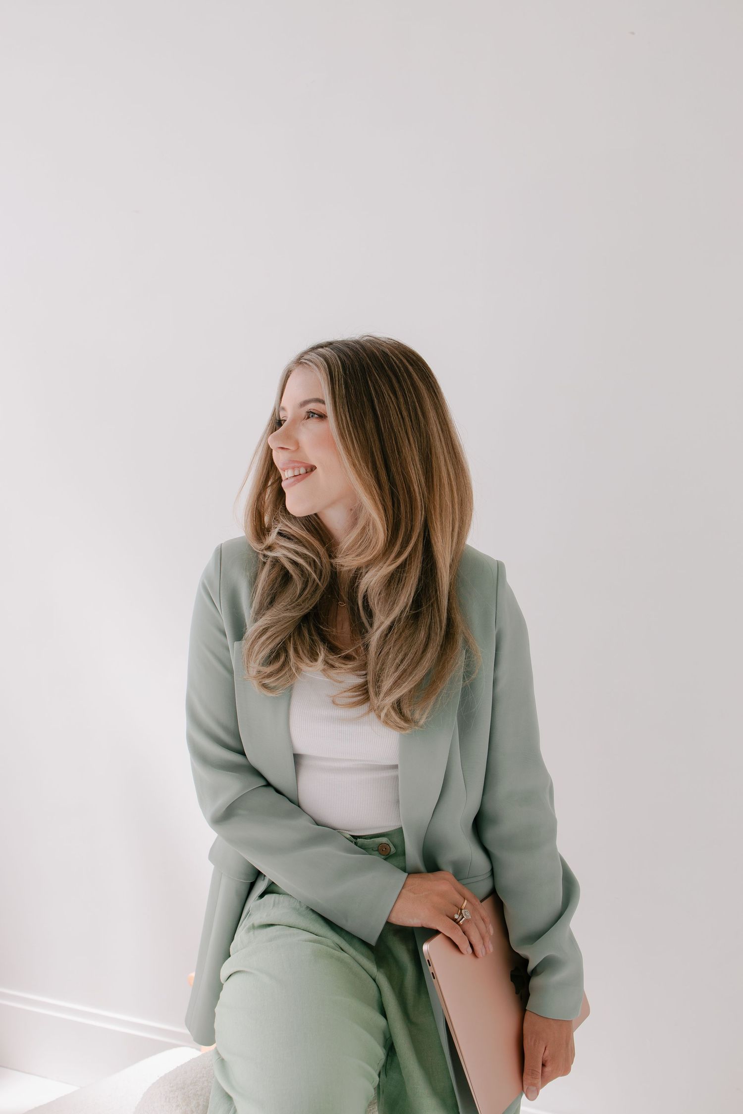 A person wearing a sage green blazer and pants with a white top sits casually while looking to the side and smiling.