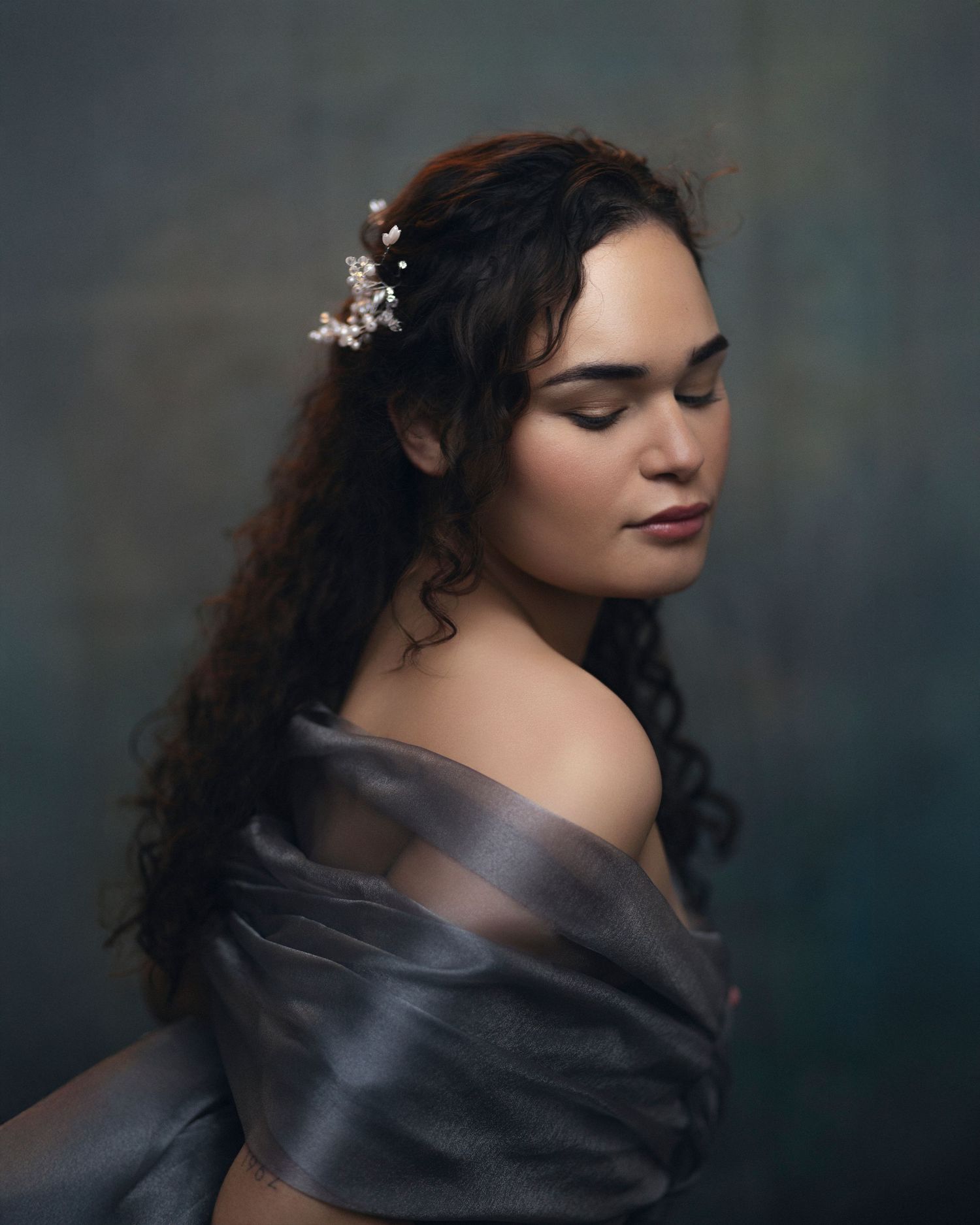 Young woman with curly hair adorned with floral pins, wearing an off-shoulder gray gown, posed gracefully against a dark backdrop.