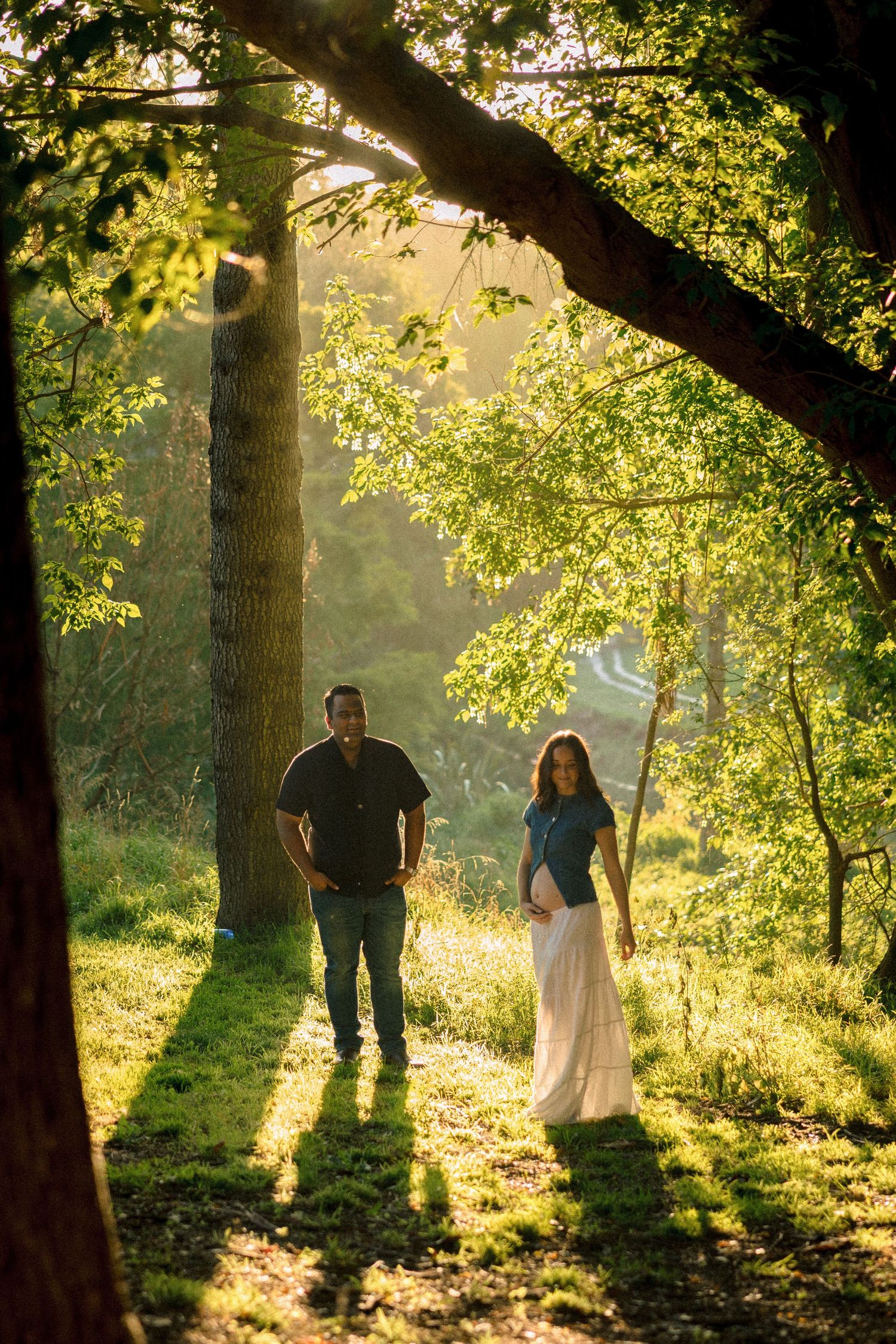 Couple walking through sunlit forest with golden light streaming through tree branches in romantic woodland setting.