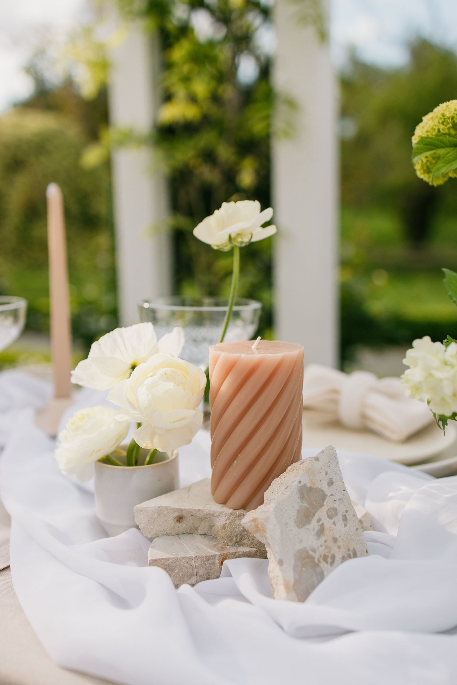 Elegant table setting with white flowers, striped peach candle, and natural stone decor on flowing white fabric.