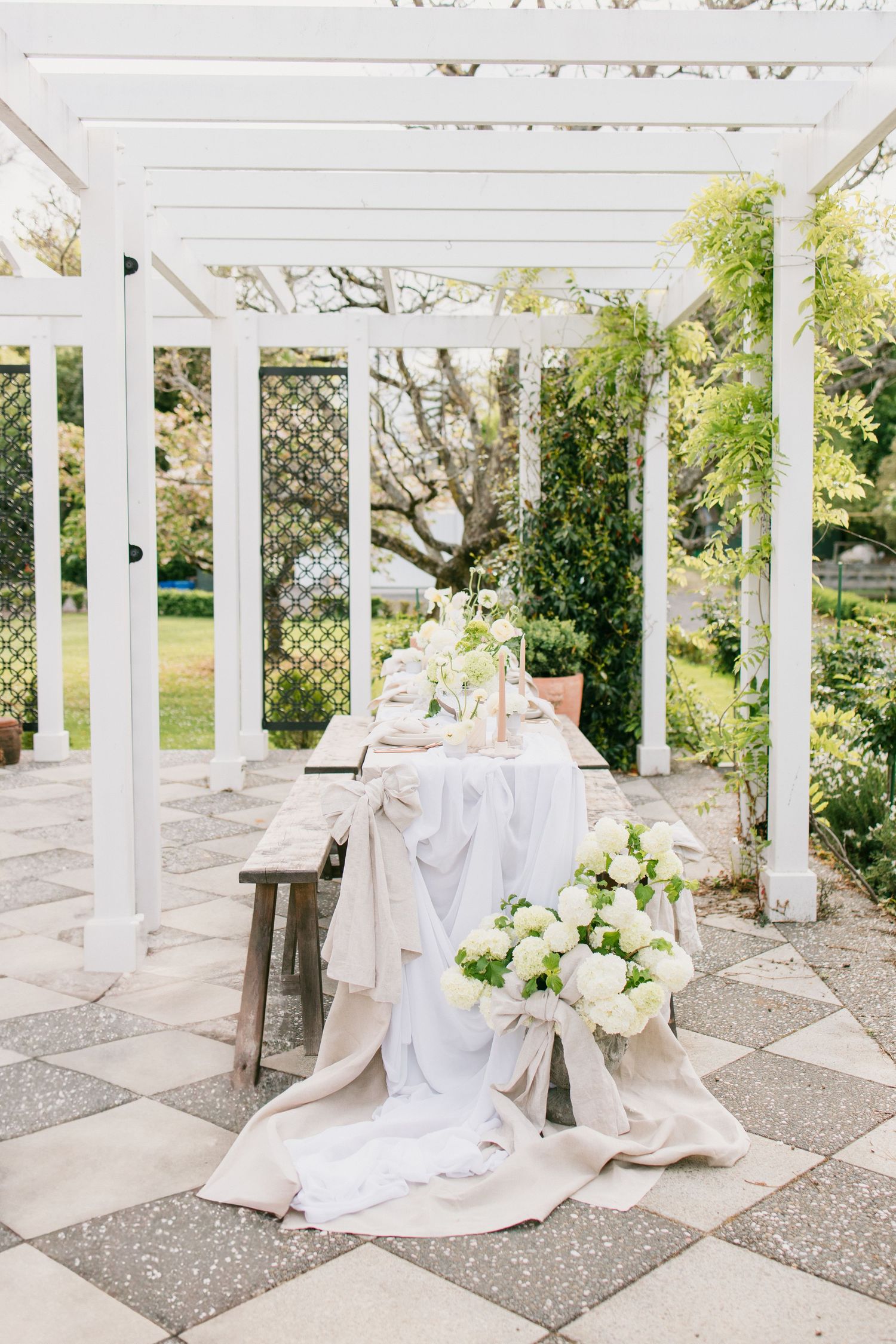 Elegant white pergola with romantic table setting and draped fabric on checkered stone patio with fresh floral arrangements.
