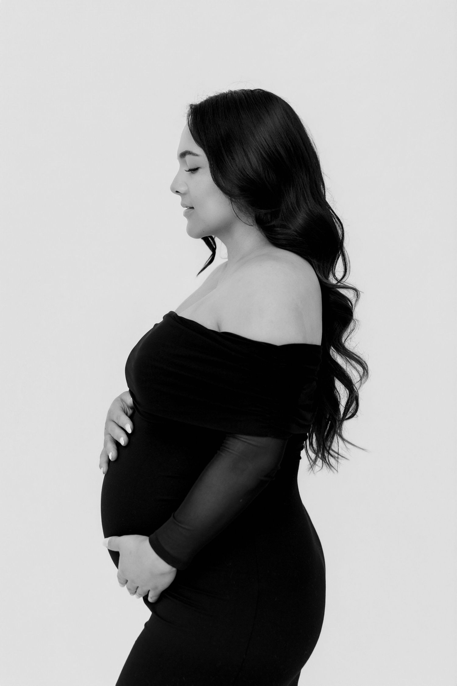 Black and white maternity photo of a pregnant woman in an off-shoulder black gown, cradling her baby bump in profile.