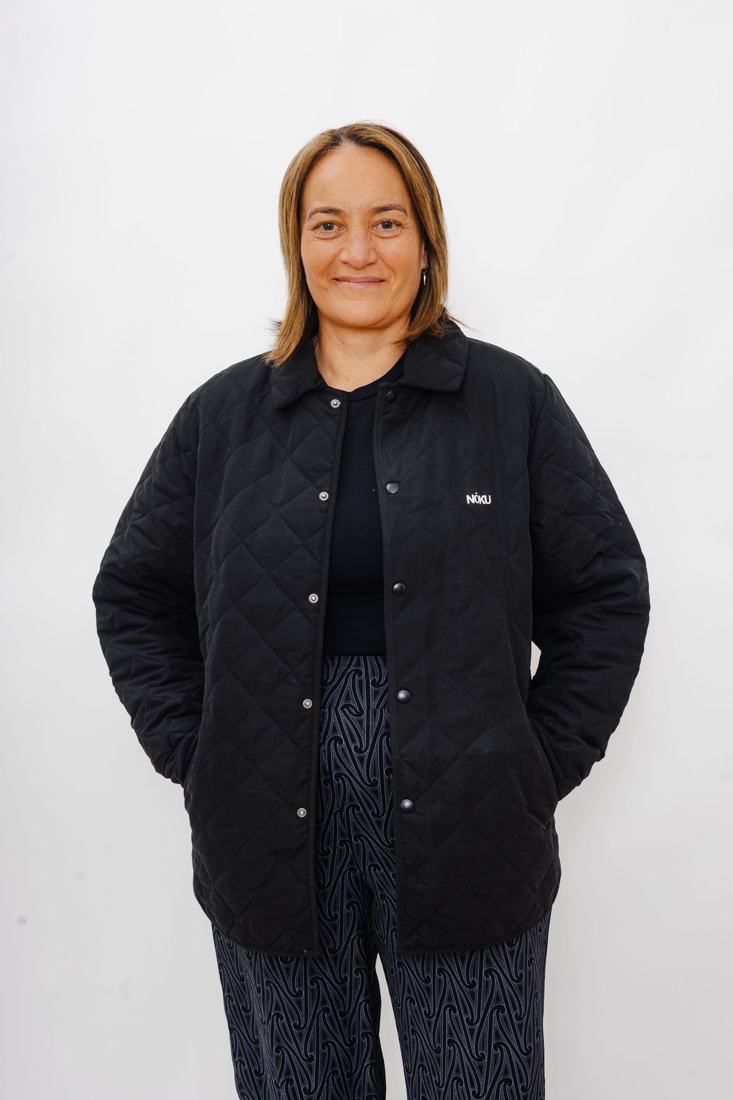 Someone wearing a black quilted jacket and patterned pants poses against a white wall.