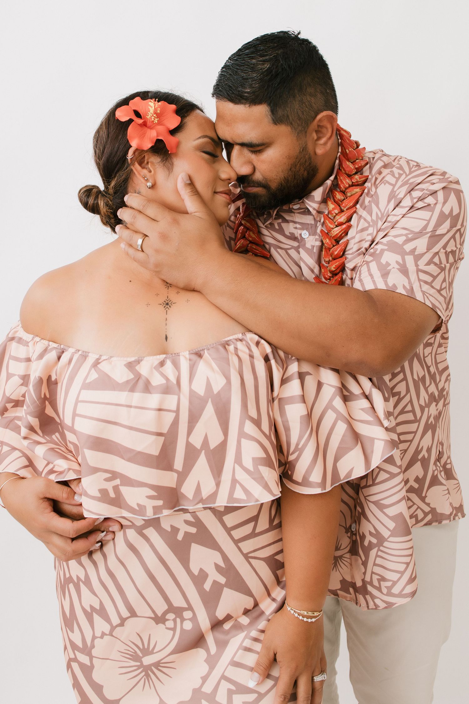 Couple in coordinating pink and beige Hawaiian print outfits embrace romantically with tropical flowers and leis.