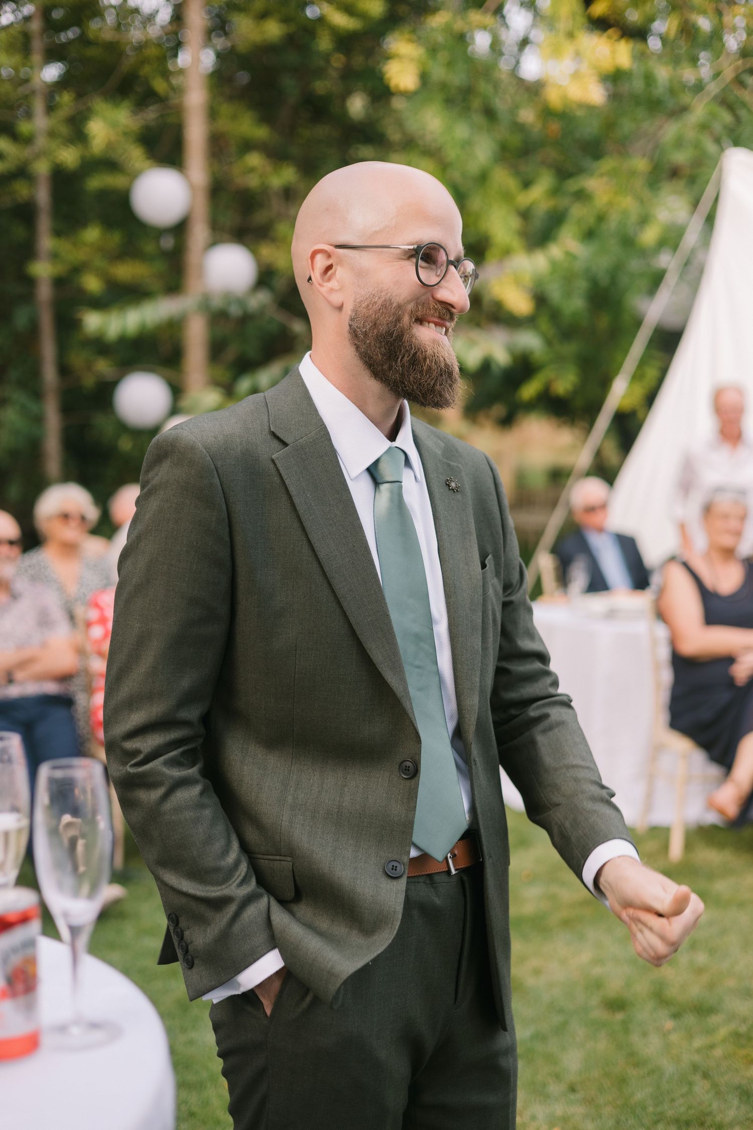 A groom in a dark suit and mint tie smiles during an outdoor garden reception.