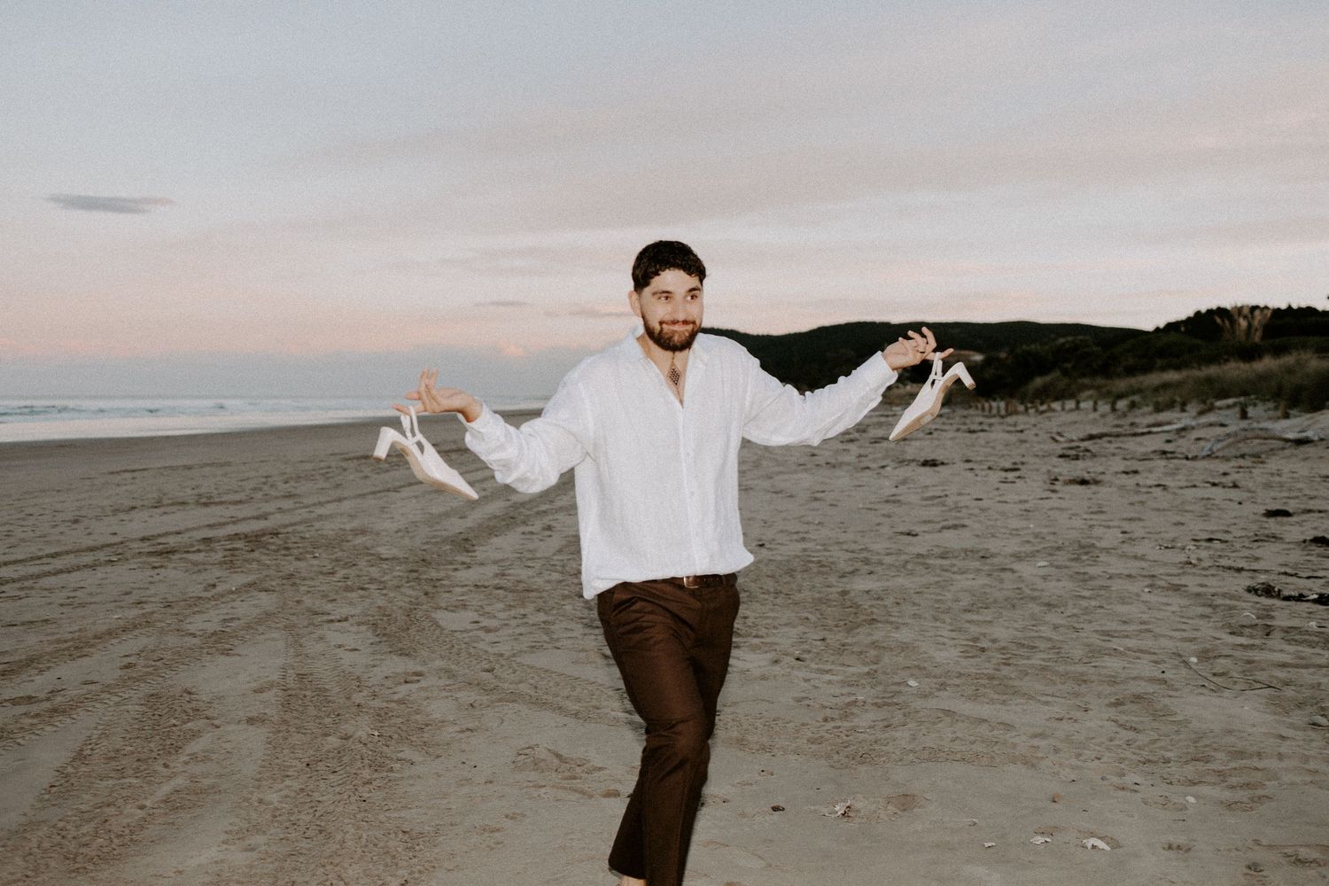 A person in white shirt and brown pants dances joyfully on a beach at dusk.