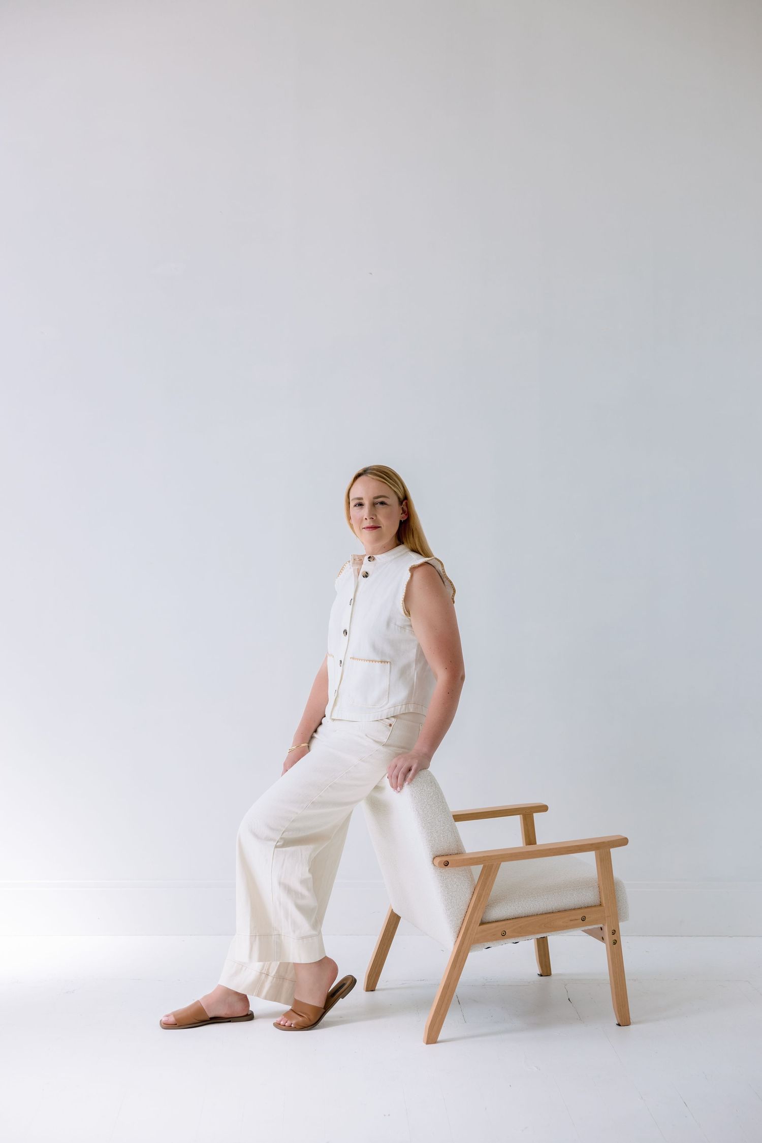 A person in an all-white outfit poses against a minimalist backdrop with a wooden chair.