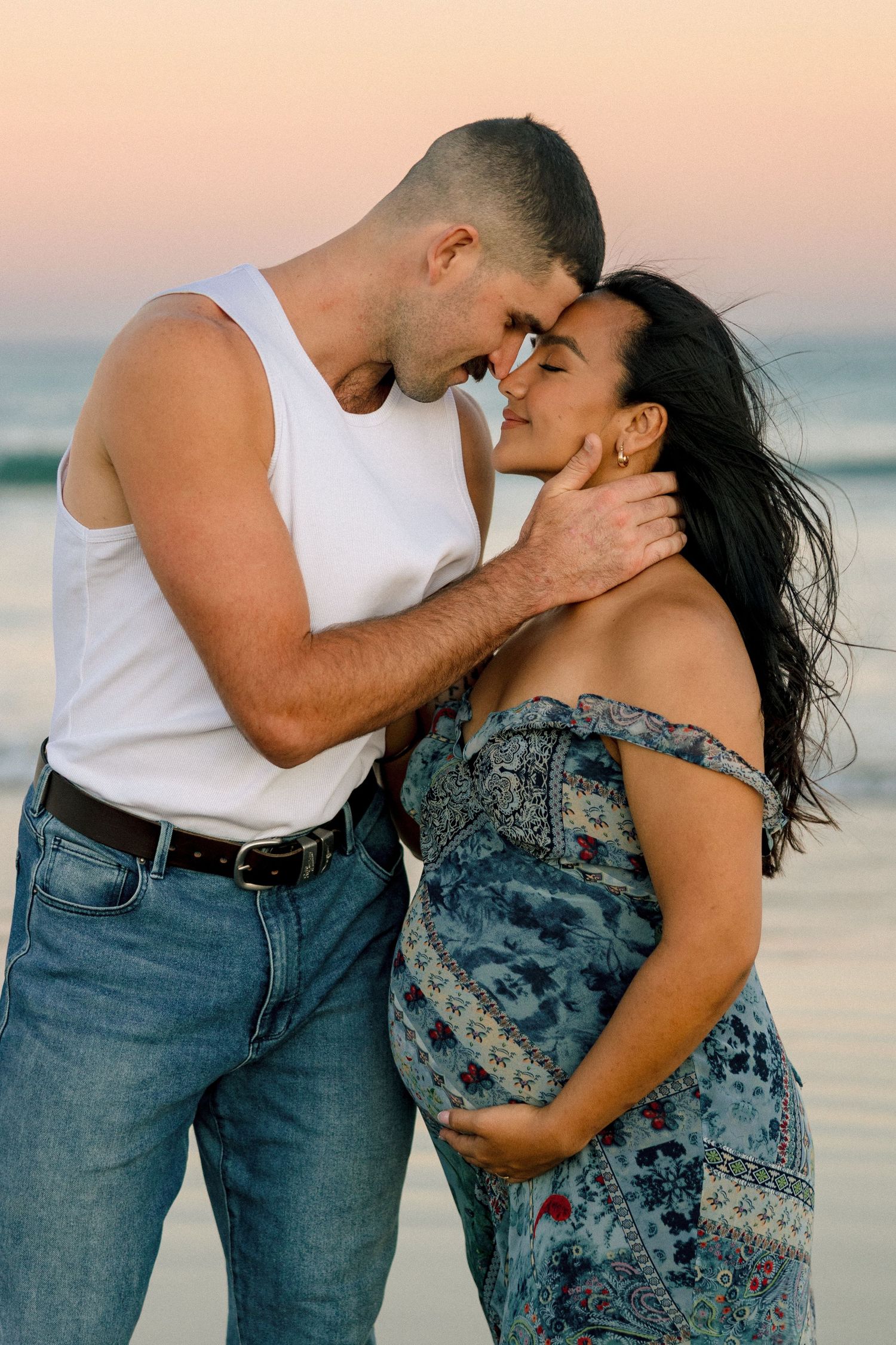 A romantic couple embracing and kissing on the beach at sunset wearing casual outfits of jeans and white tank top.