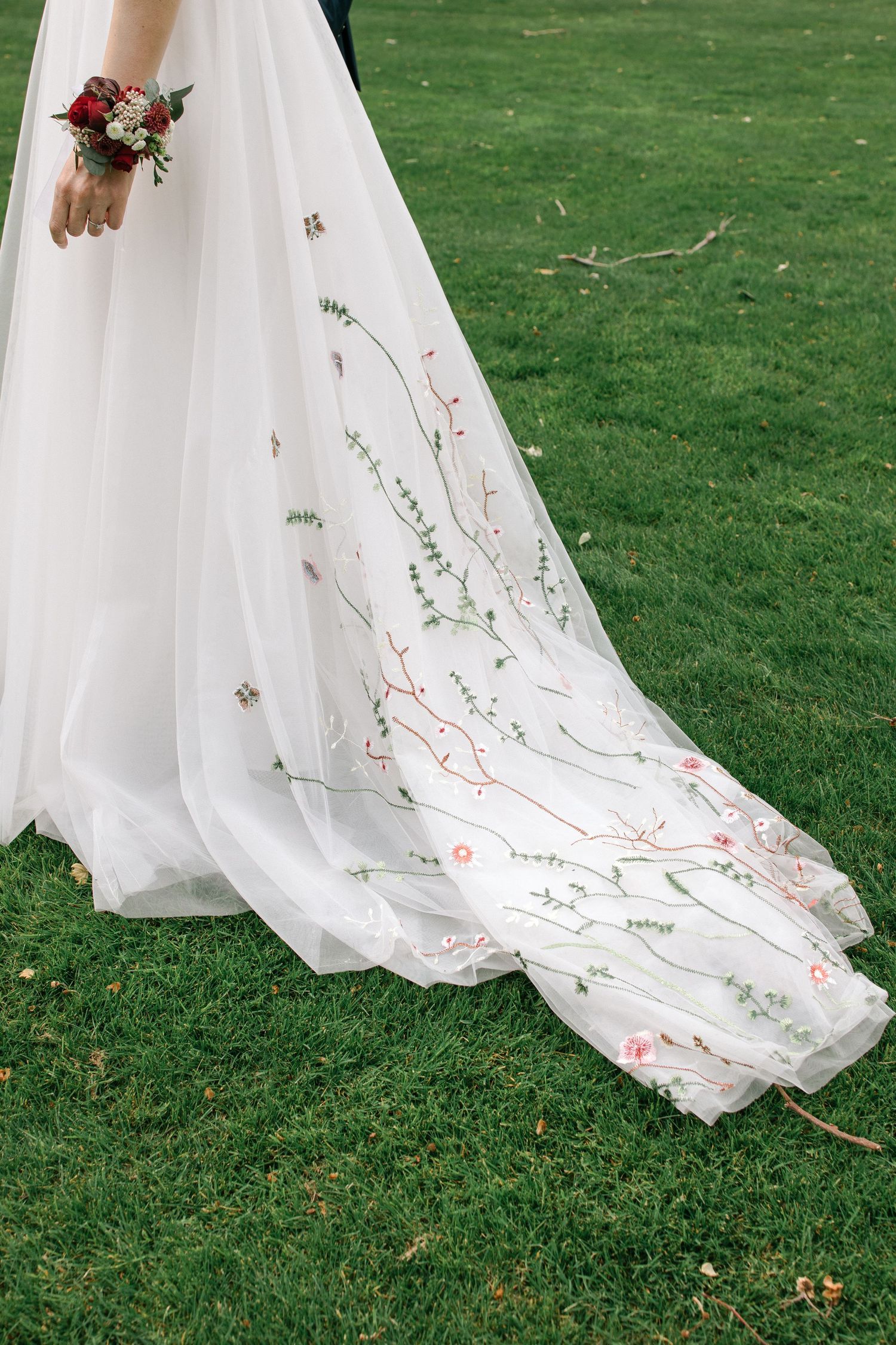 Close-up of a white wedding dress train with delicate floral embroidery trailing on green grass.