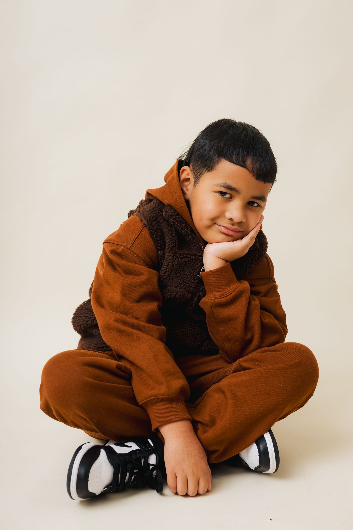 A person in a brown oversized sweatsuit and black sneakers poses casually on a white background.