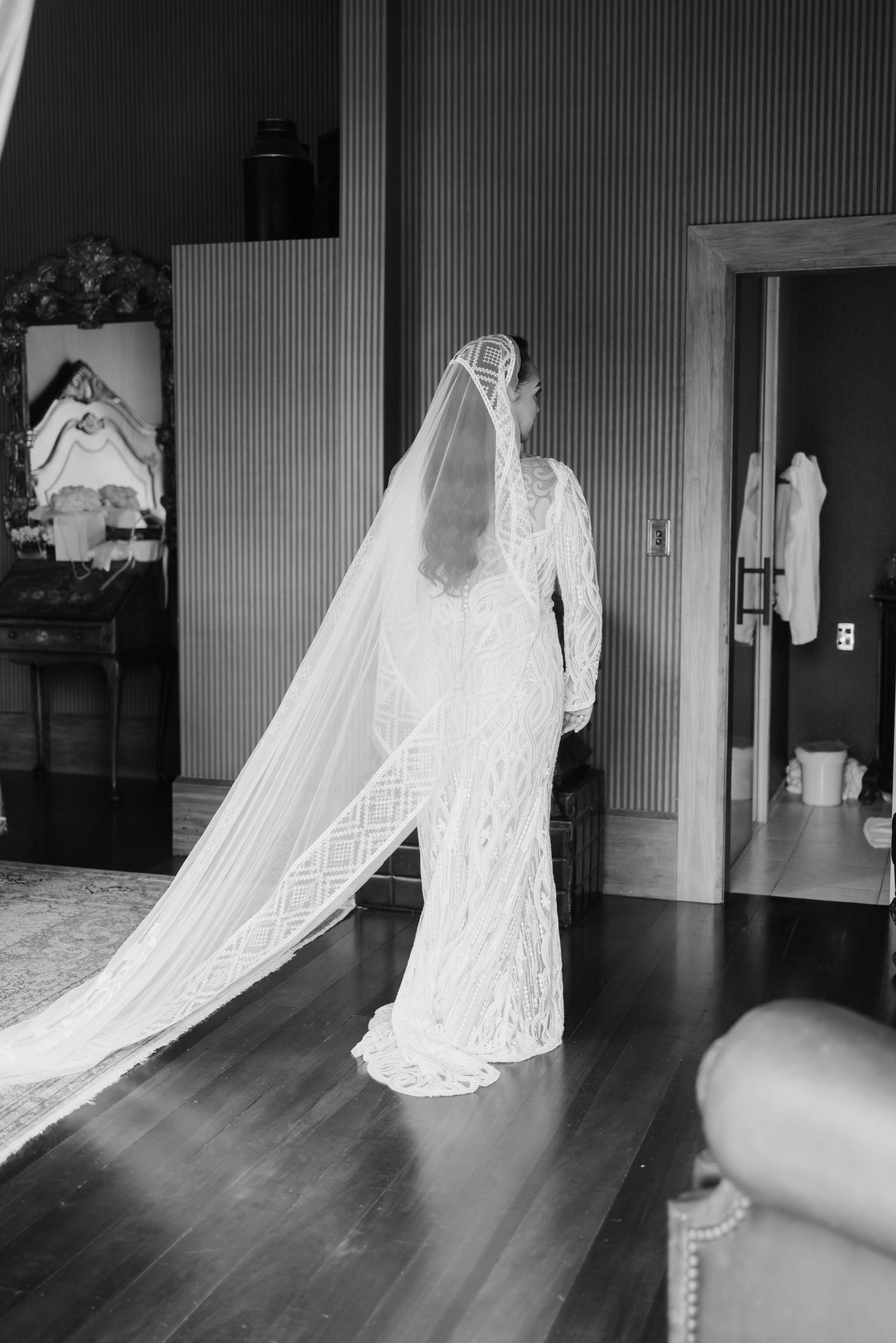 A bride in a long-sleeved lace wedding gown with a dramatic cathedral veil stands in a room with hardwood floors.