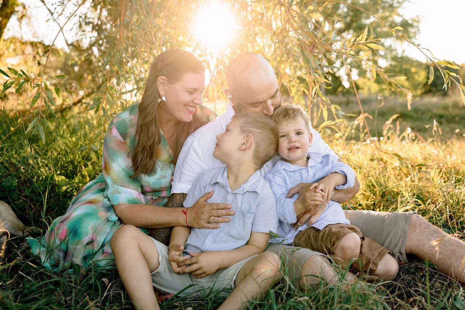 Family of four sitting together on grass in golden sunset light, two young boys laughing with parents in outdoor portrait.