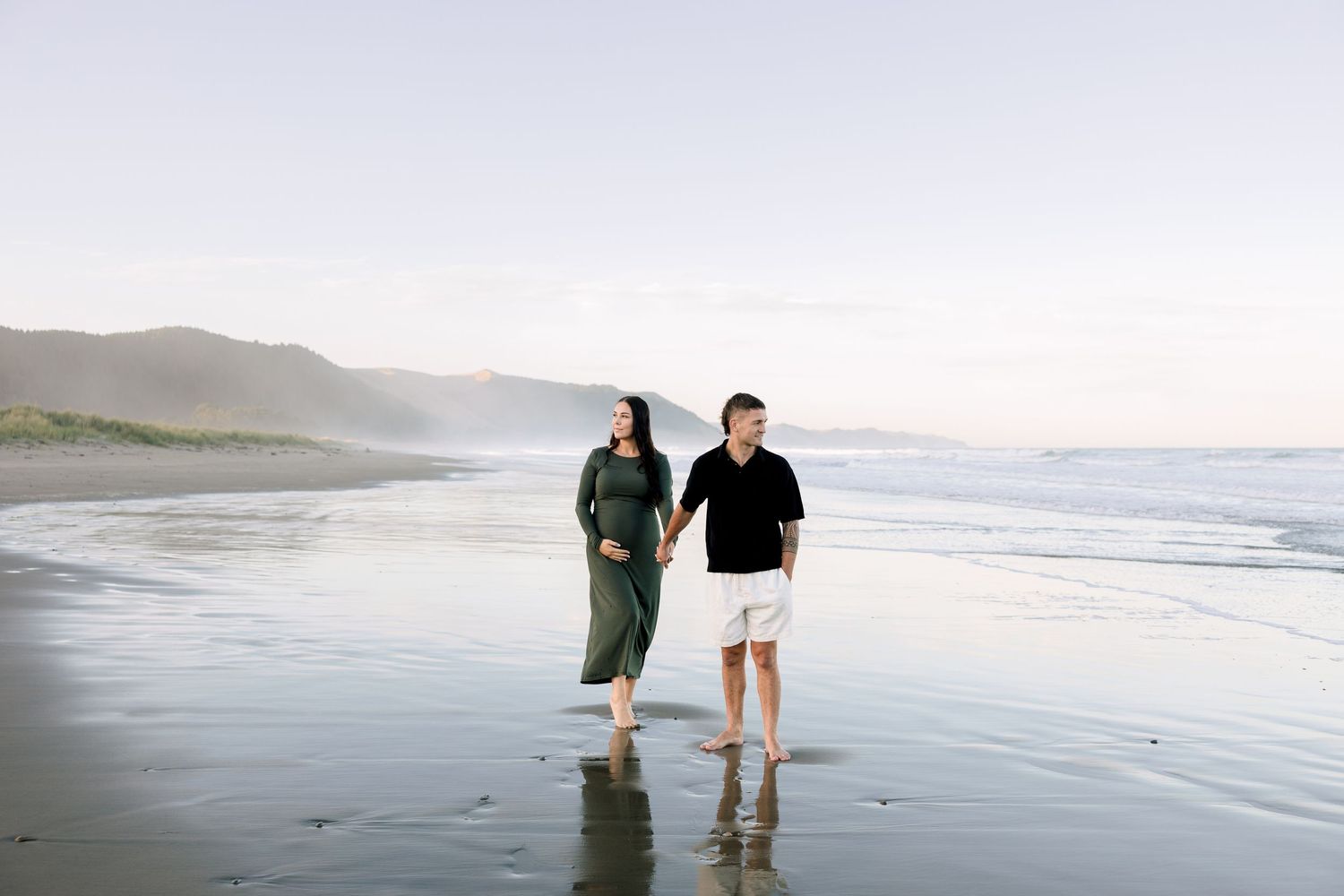 Couple walking hand in hand on a reflective beach at sunset with misty mountains in the background.