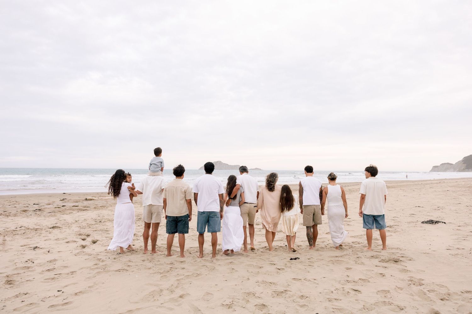 Large family group standing together on a sandy beach, viewed from behind, facing the ocean waves.