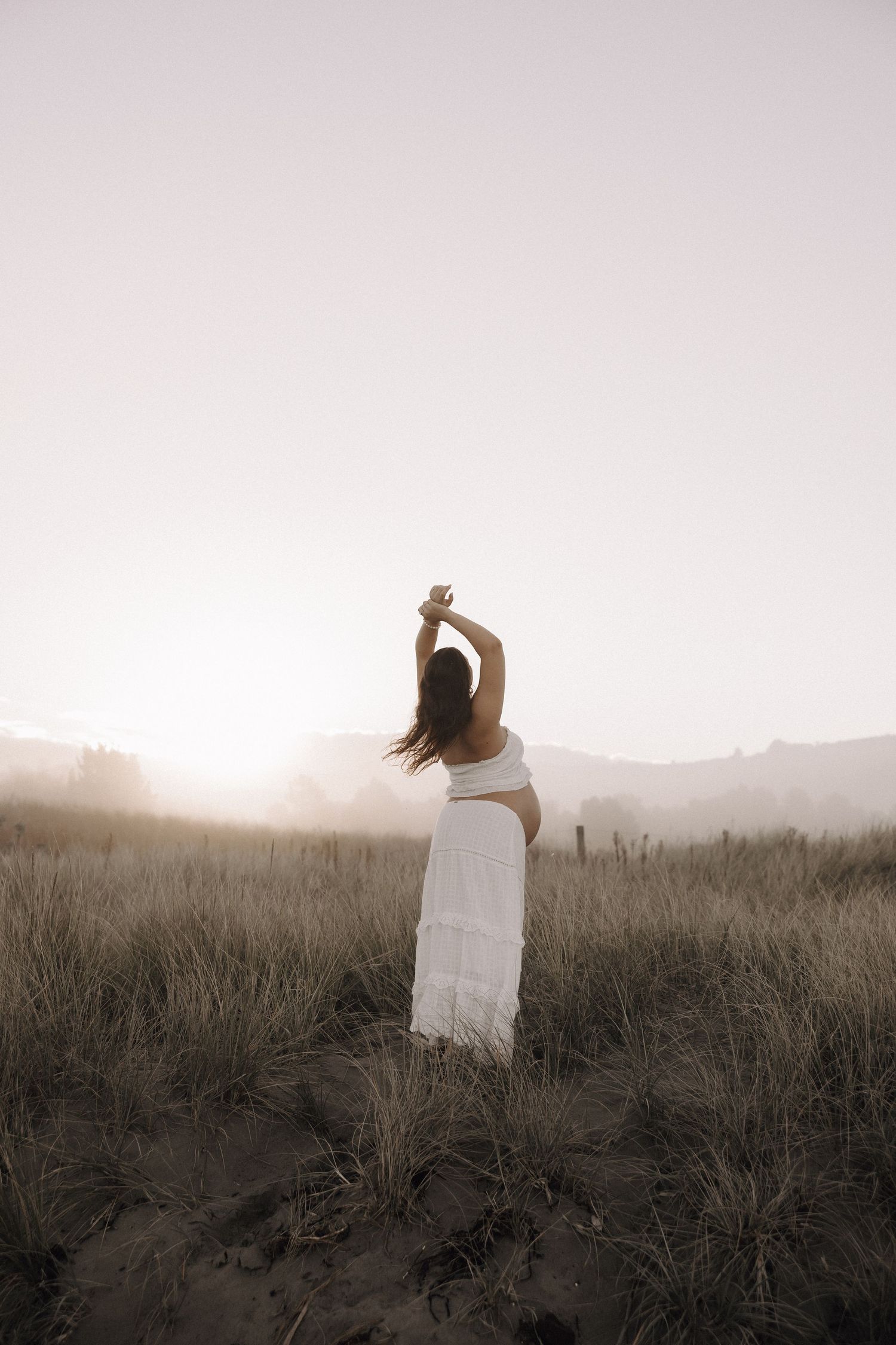 Woman in white dress standing in misty field at sunset with arms raised.