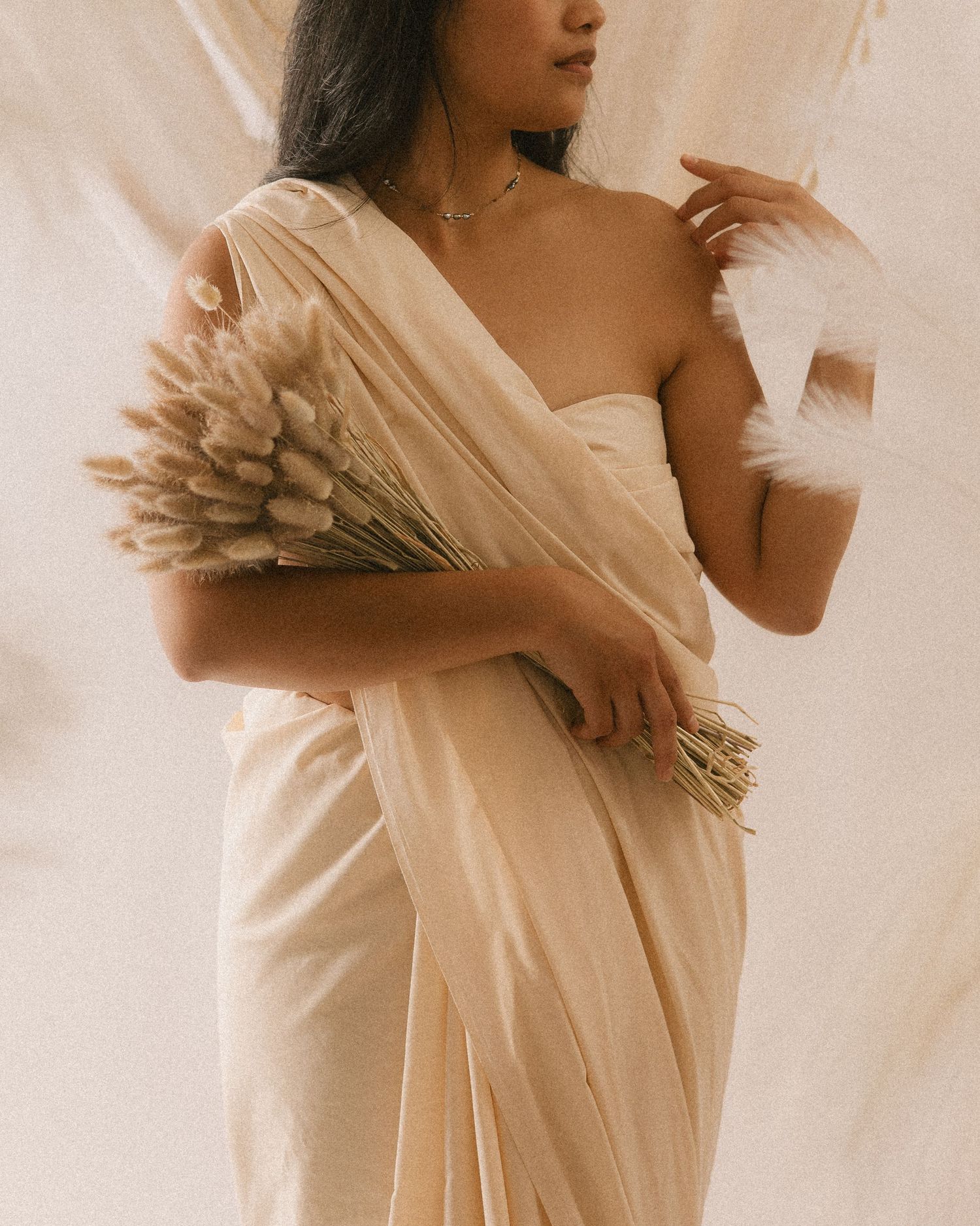 A woman in a cream satin saree holds dried pampas grass, posing gracefully against a soft beige draped backdrop.