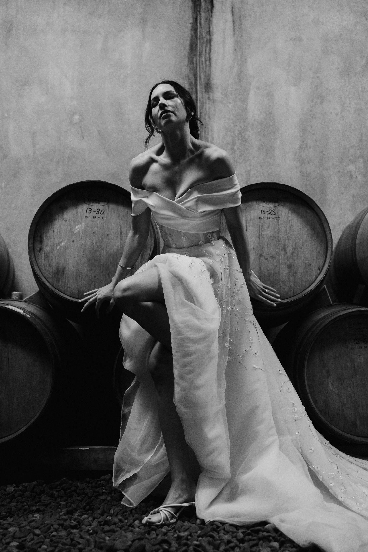 Black and white artistic bridal portrait with off-shoulder gown posed against wine barrels in rustic cellar.