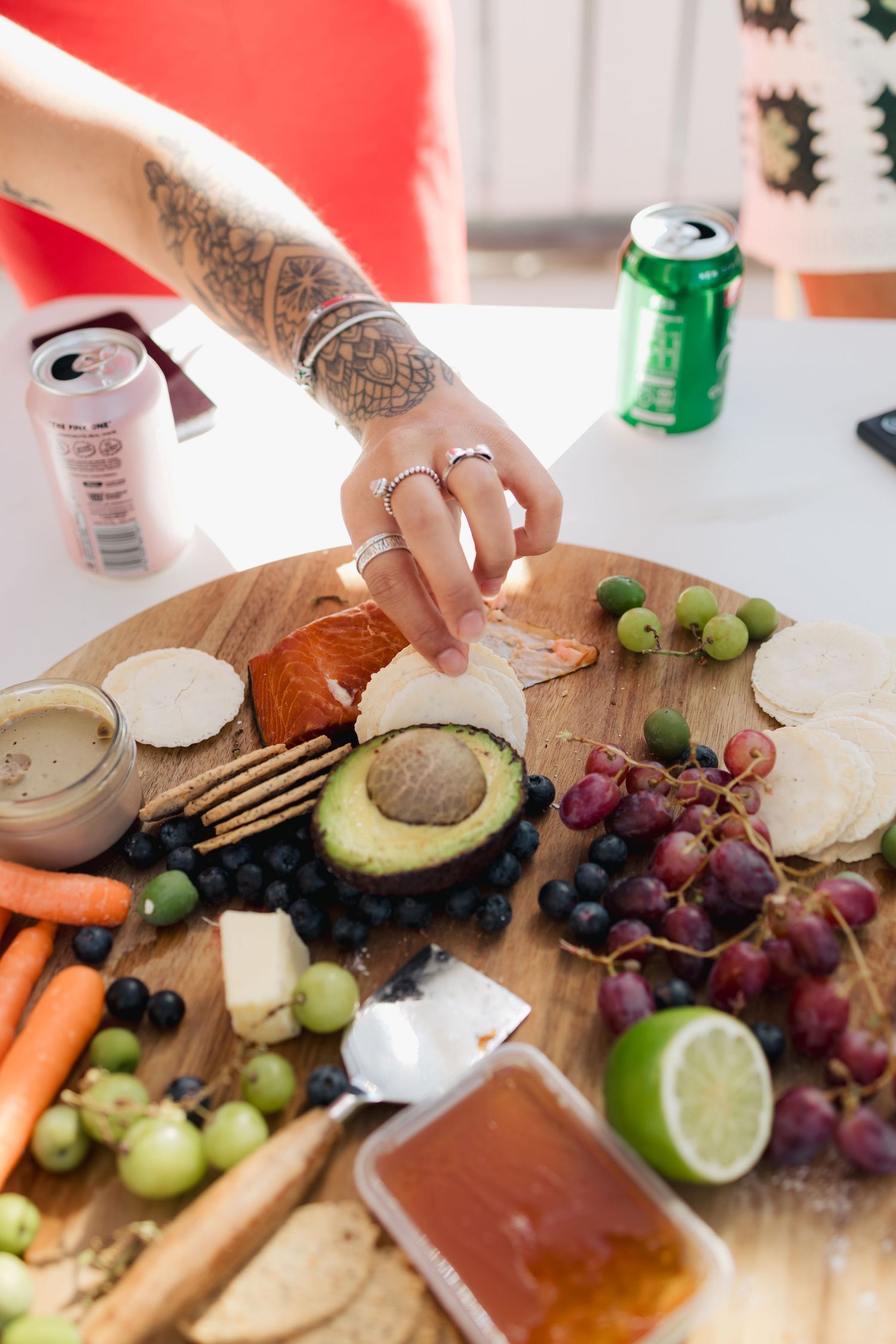 Tattooed hand reaching for food on charcuterie board with fresh fruits and vegetables.