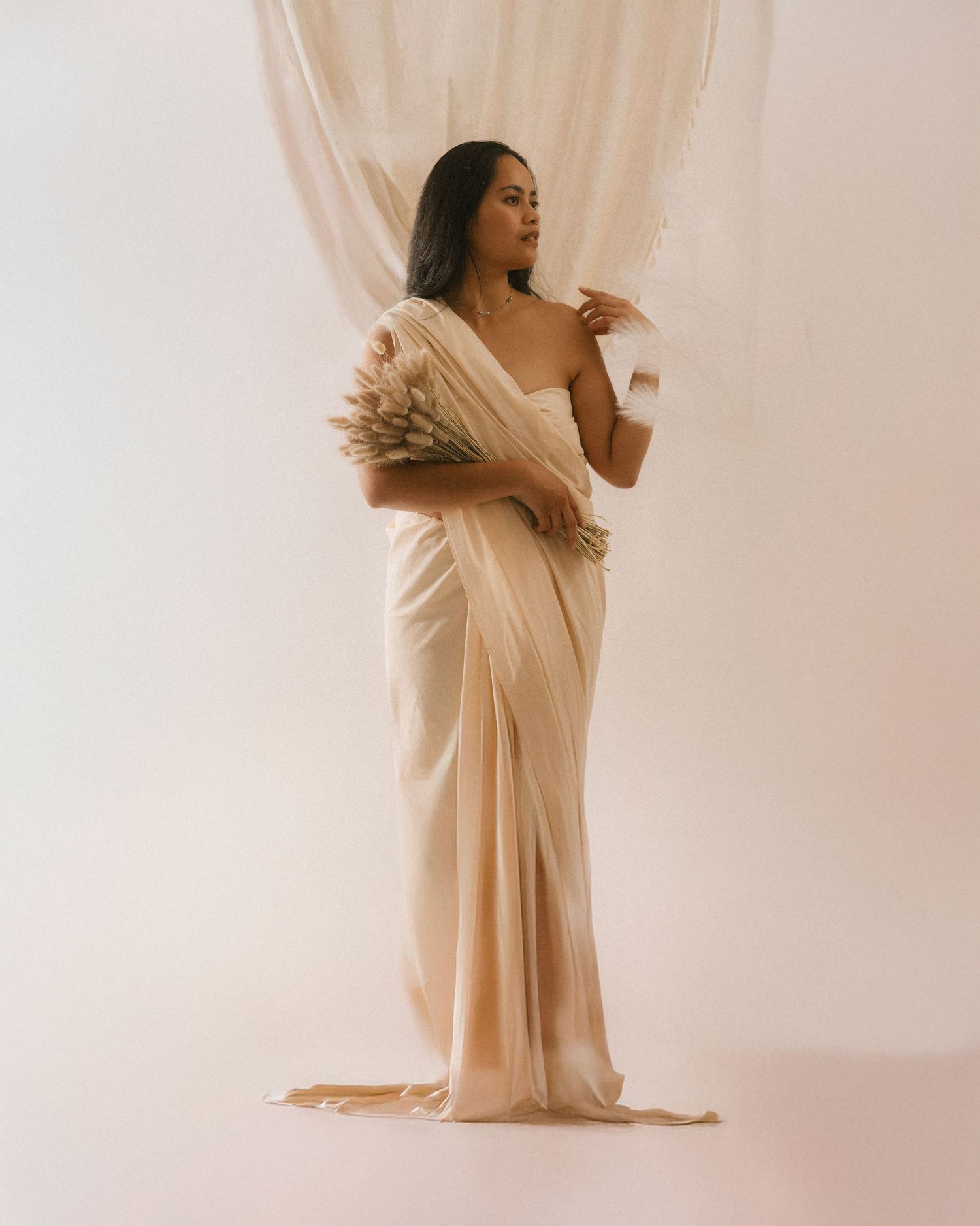 Elegant figure in flowing cream-colored dress poses gracefully against sheer white curtain background in artistic portrait.