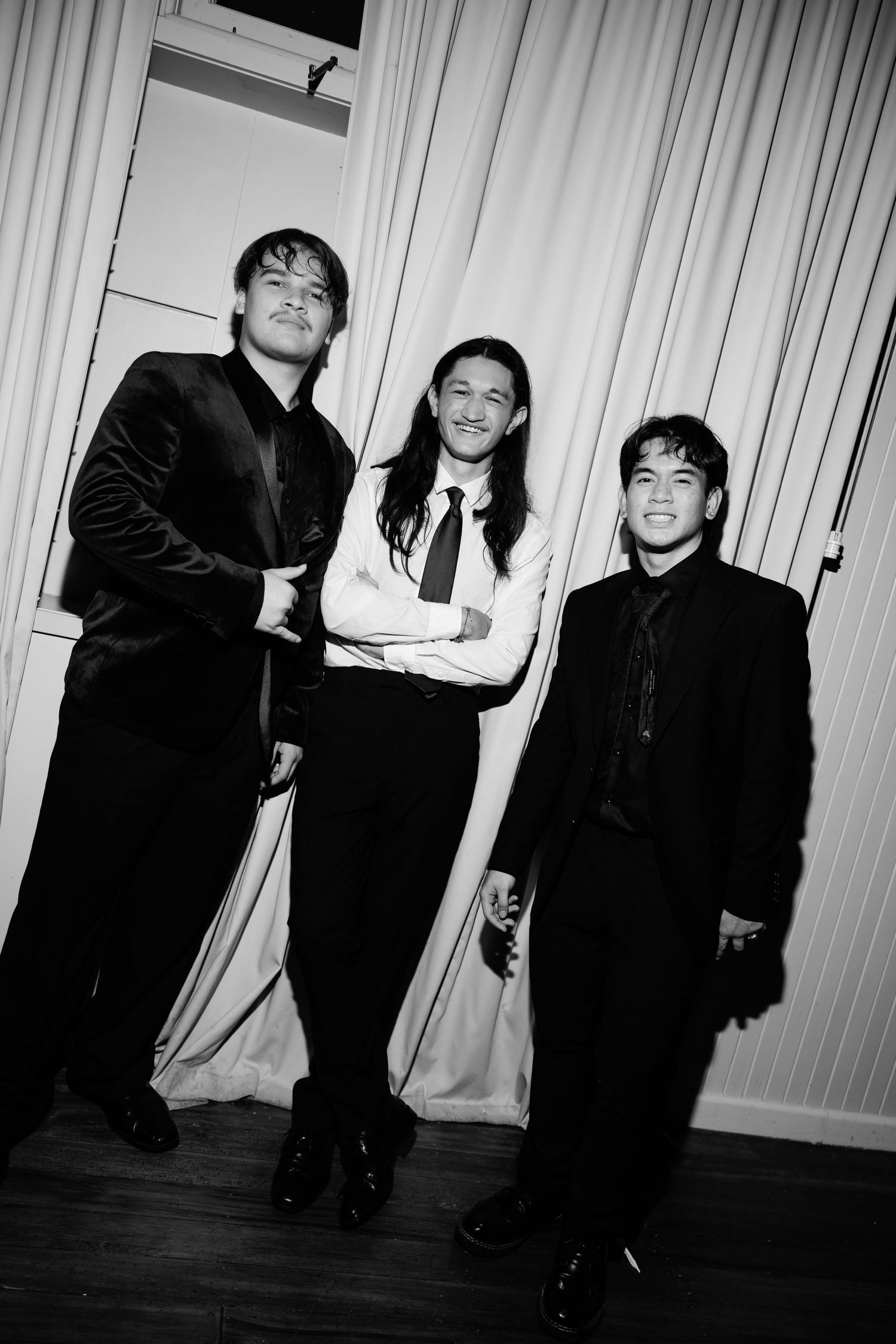 Three young men in formal black suits pose together smiling in a black and white photo.