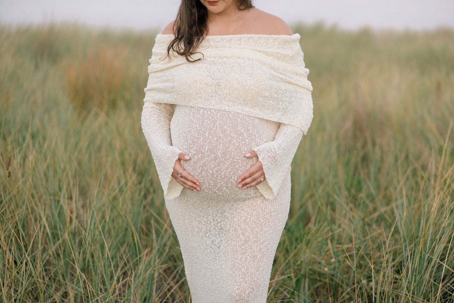 Pregnant woman in white off-shoulder sequin gown cradling baby bump in a lush green grass field.