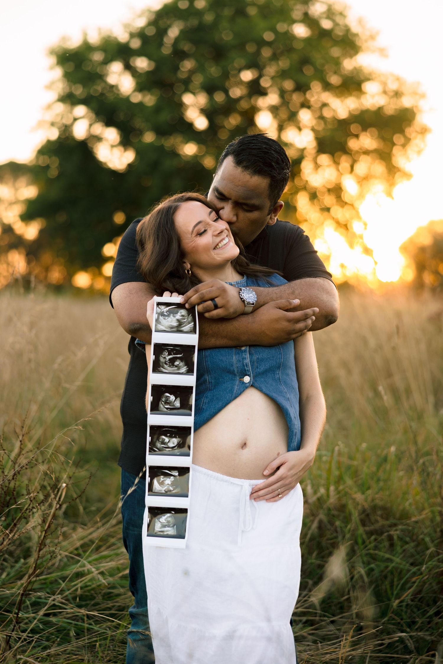 A couple embraces in a golden sunset field while holding ultrasound photos showing their expected baby.