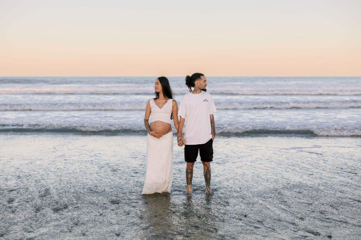 Pregnant couple holding hands at ocean's edge during pastel sunset, woman in white dress.