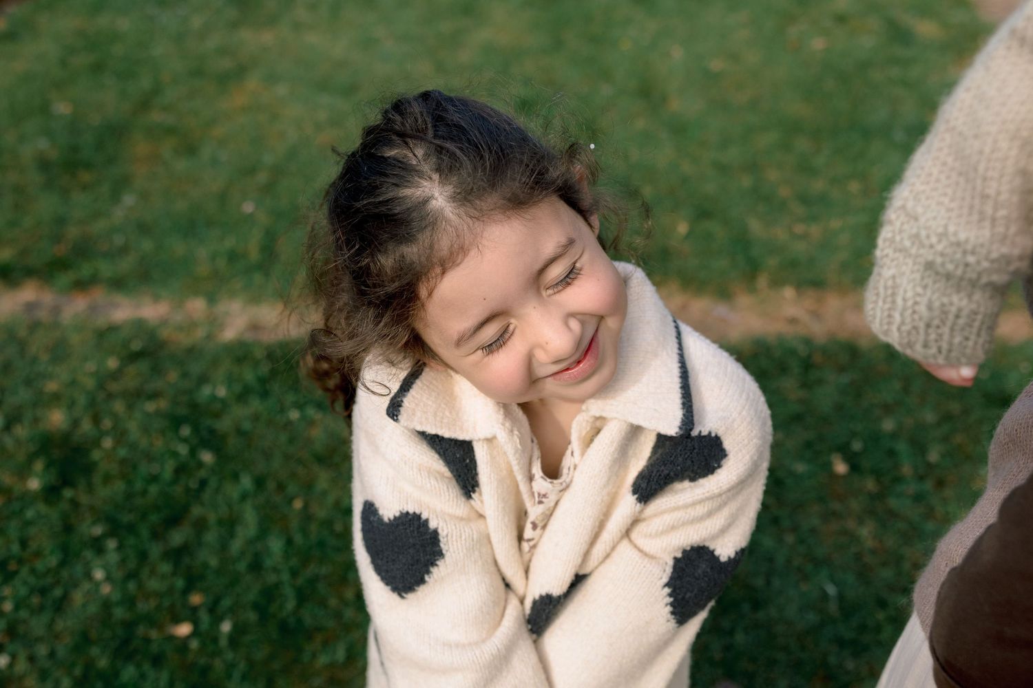 Smiling young girl in cream knit heart cardigan sweater standing on green grass outdoors.