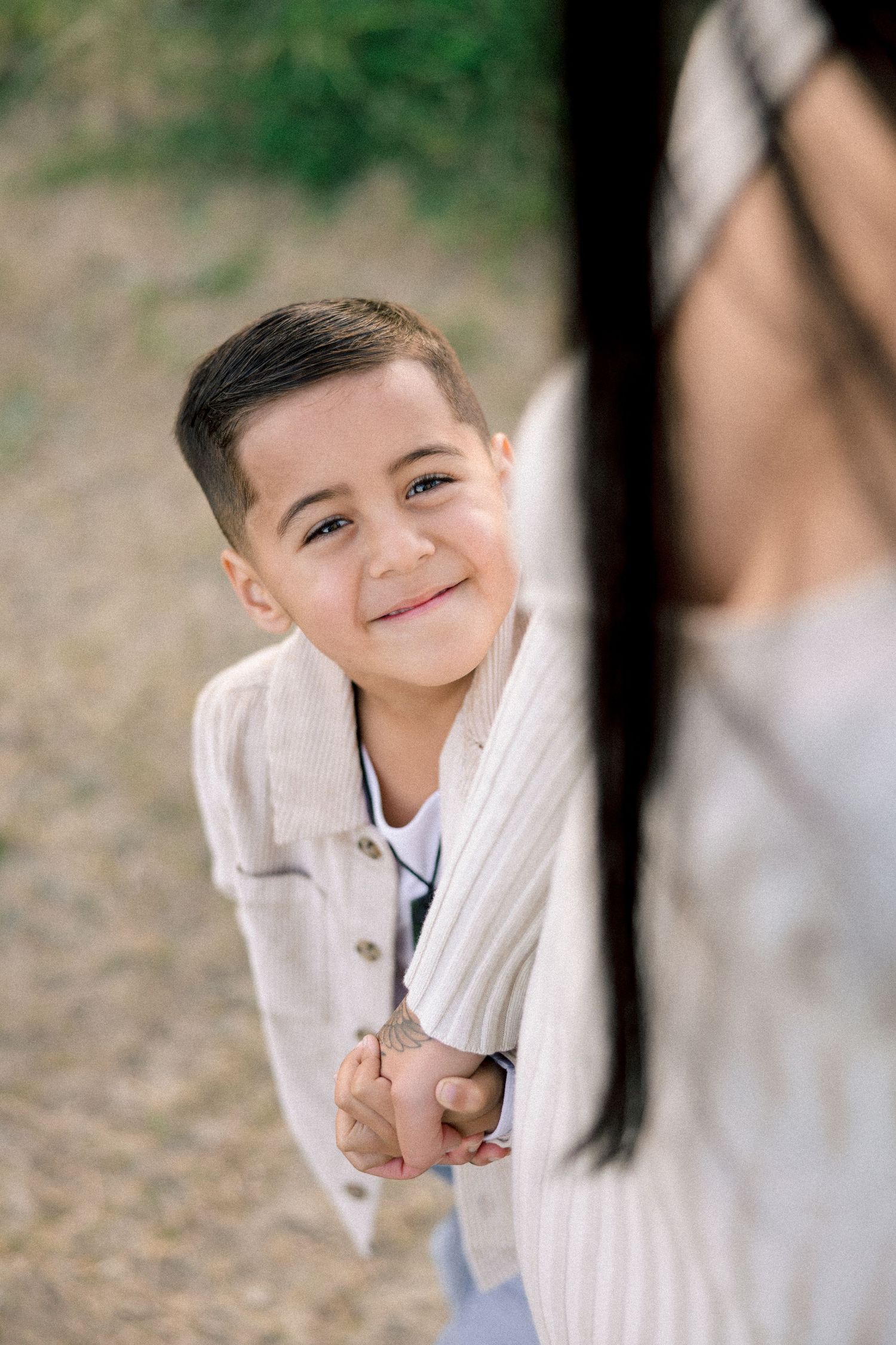 Smiling young boy in beige jacket holds hands with dark-haired woman outdoors, looking up with joy.