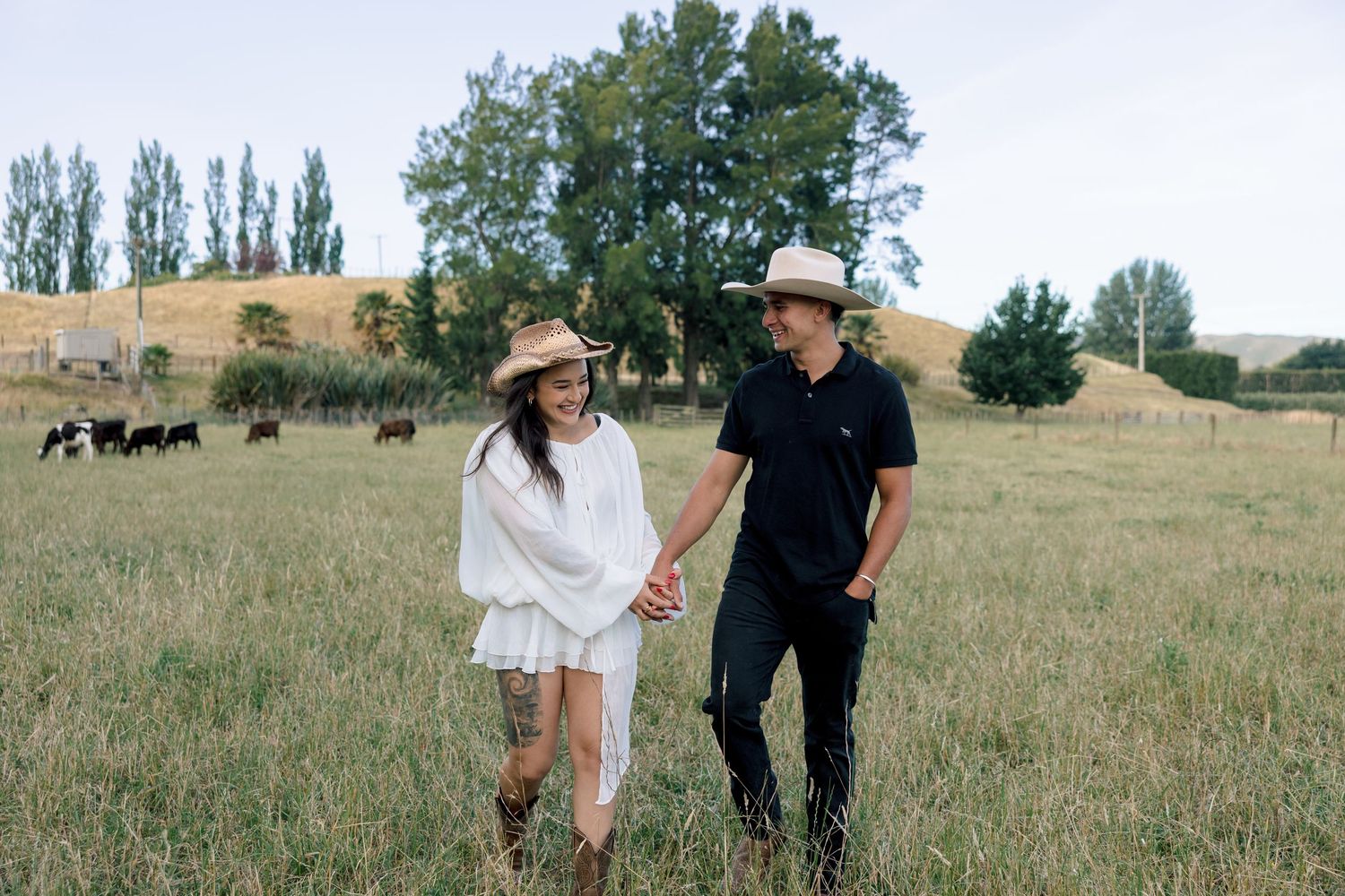 A romantic couple walks through a grassy field with trees in the background on a sunny day.