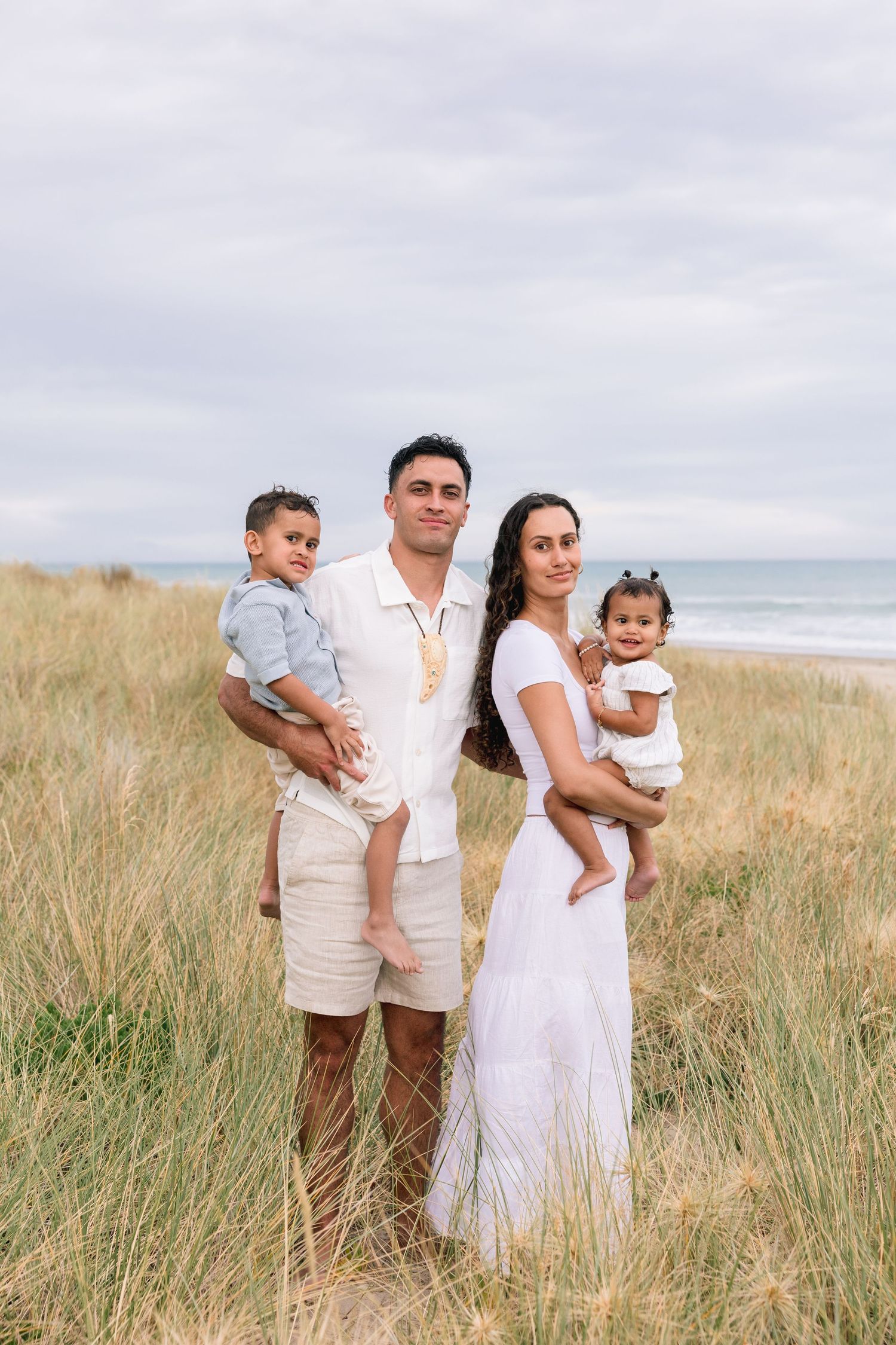A family in white and neutral clothing poses together among tall beach grass with the ocean visible in the background.