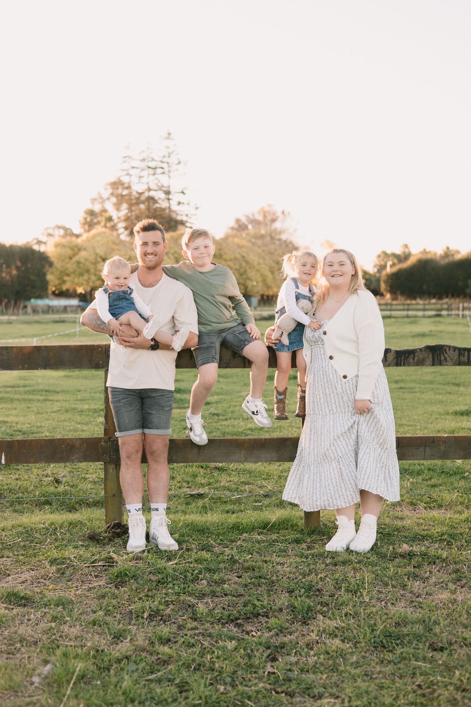 Parents and children posing together against a rustic wooden fence in a grassy field at sunset.