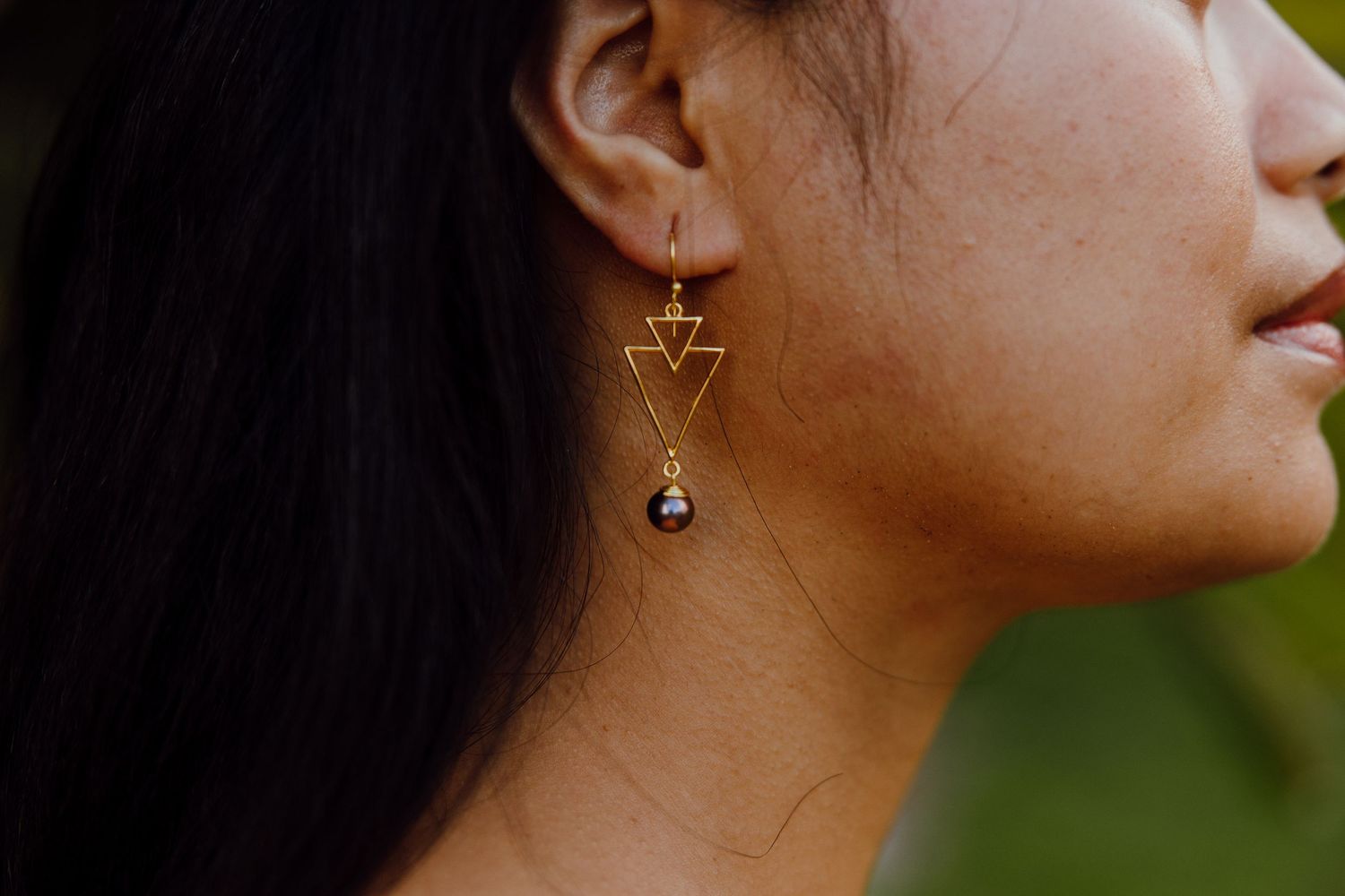 Close-up of a geometric gold triangle dangle earring with dark stones worn against long black hair.