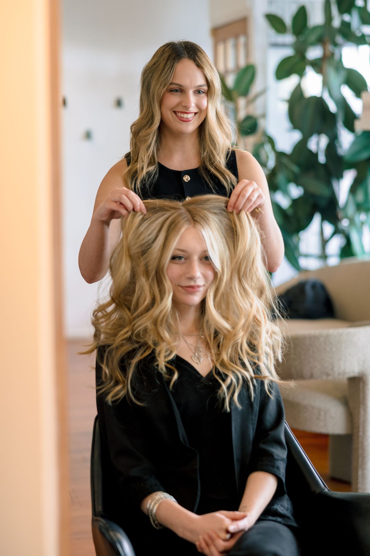 Smiling blonde hairstylist styling curly blonde hair on seated client in modern salon.