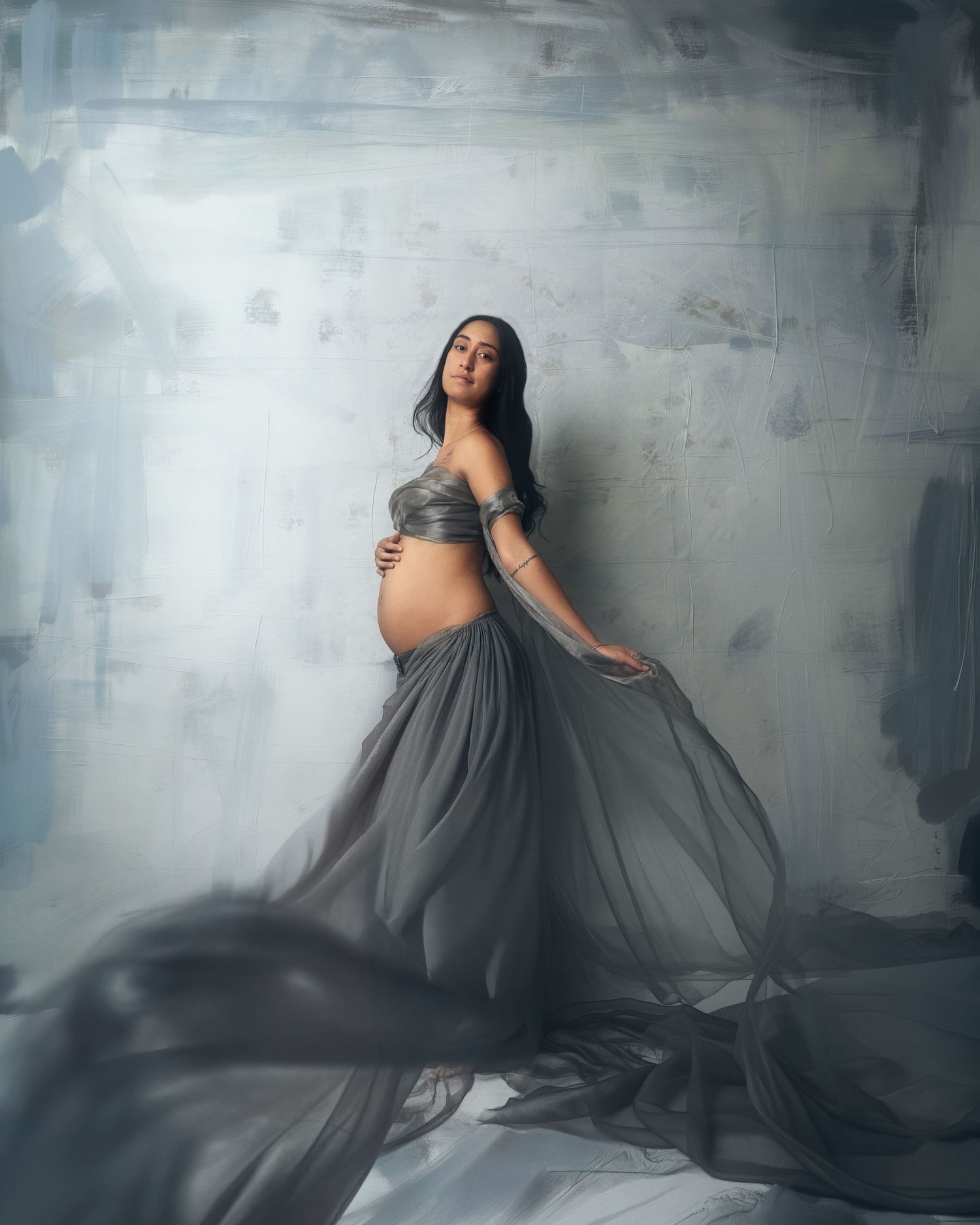 A flowing gray maternity gown creates ethereal movement against a textured concrete wall in an artistic photo series.