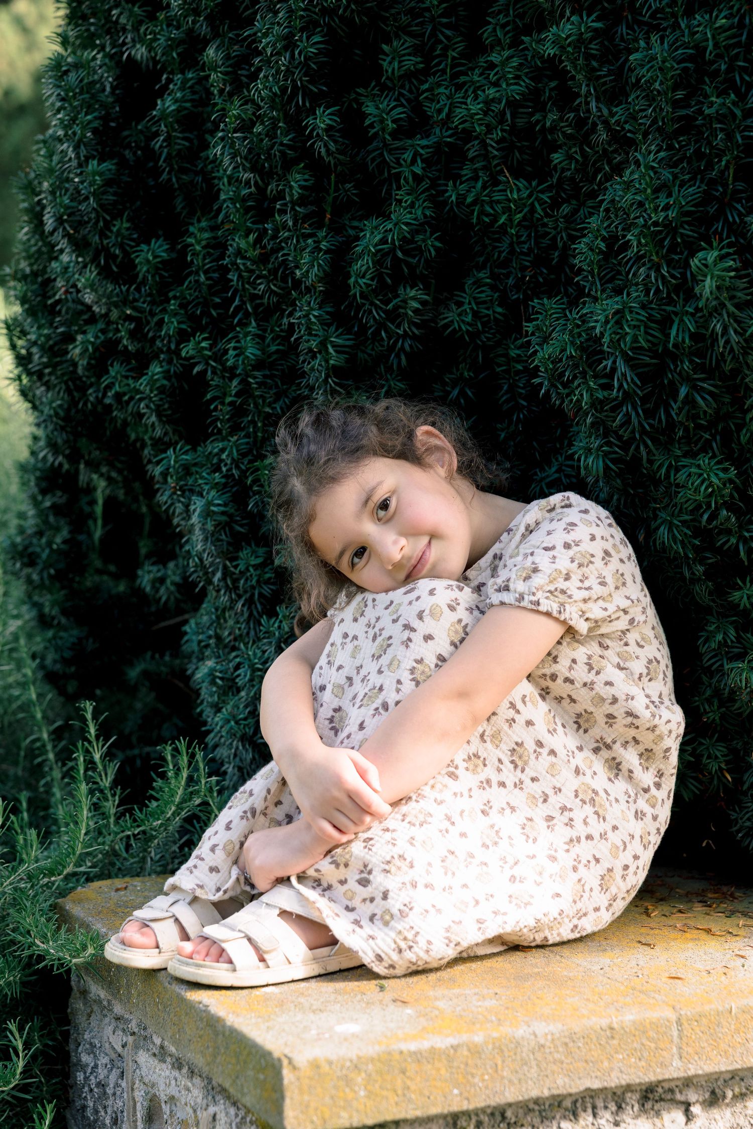 A young girl in a floral dress sits on a stone ledge hugging her knees, smiling in front of a lush evergreen shrub.
