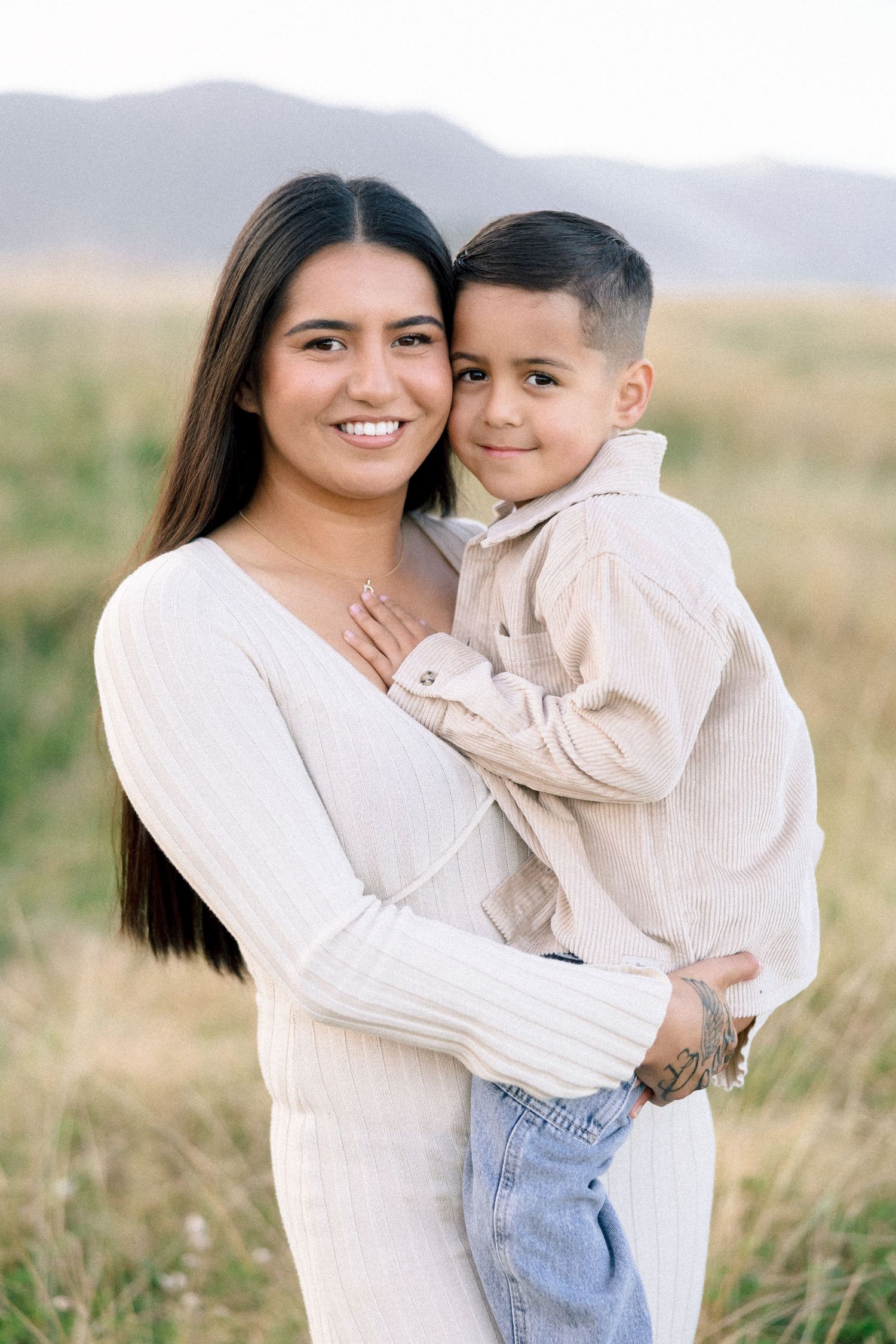 A smiling woman in white holds a young boy in a beige shirt in a grassy field.