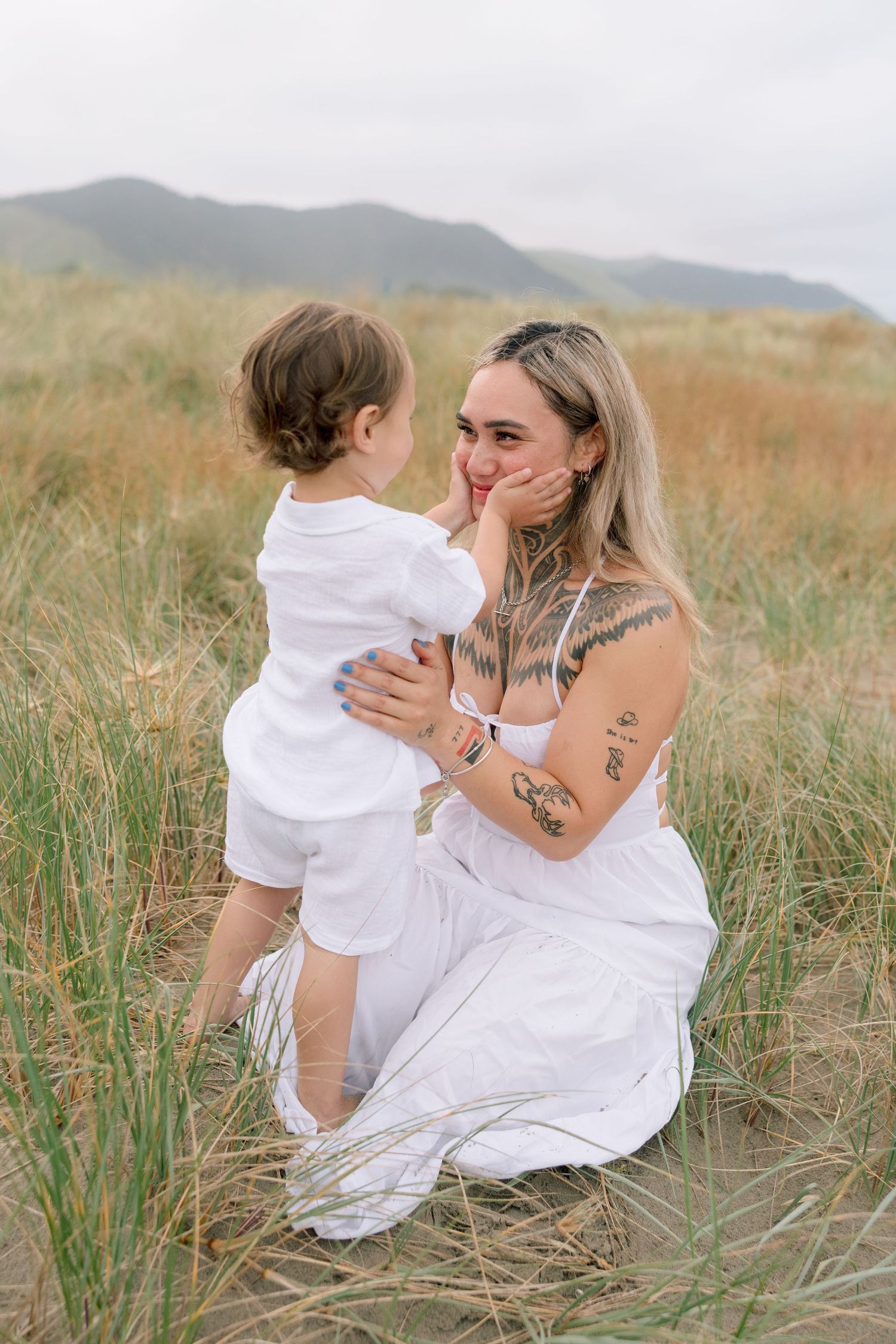 A tender moment shared between two people in white dresses sitting among tall grass with mountains in the background.