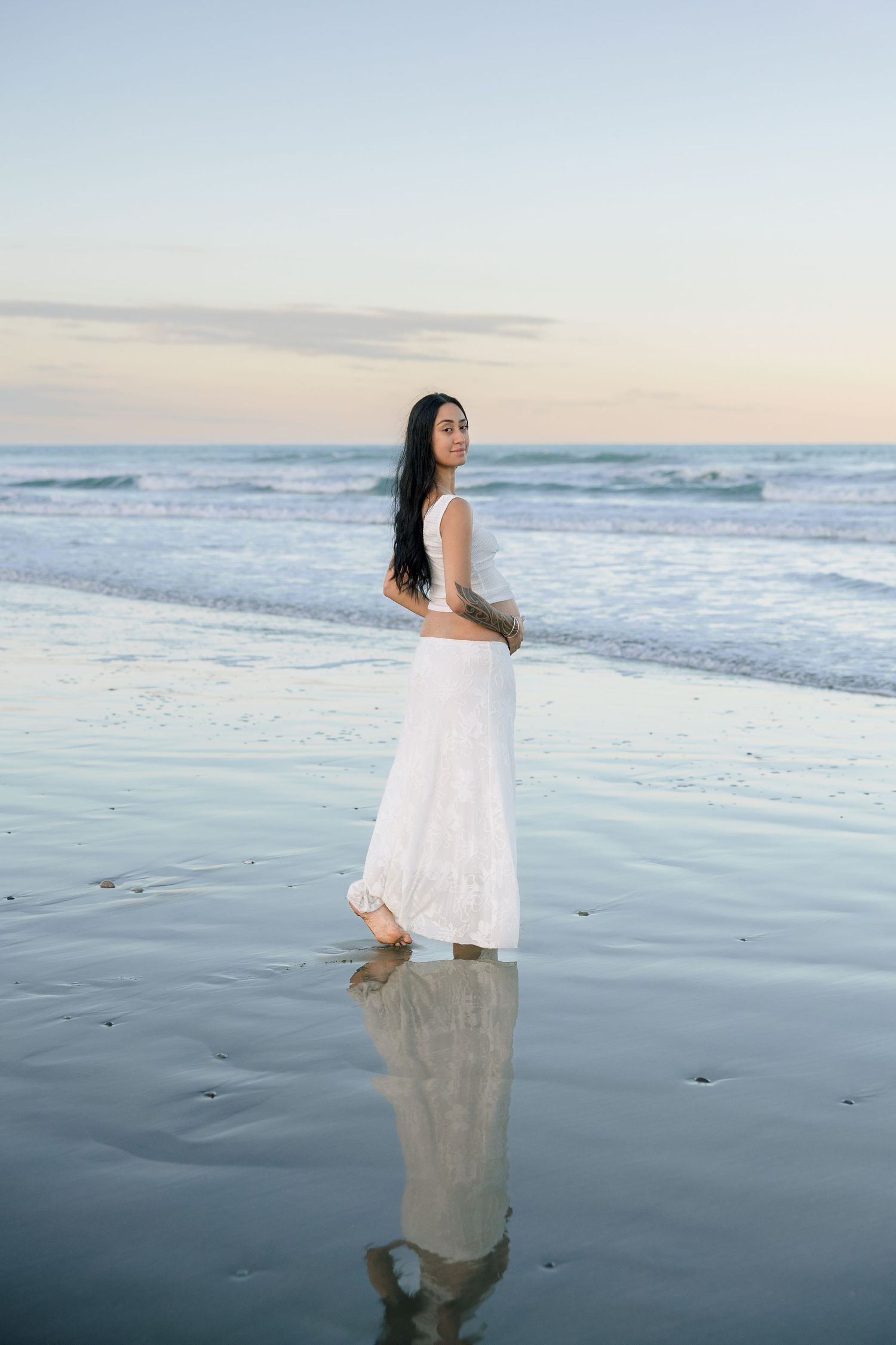 Woman in white dress standing on reflective wet beach at sunset with ocean waves behind her.