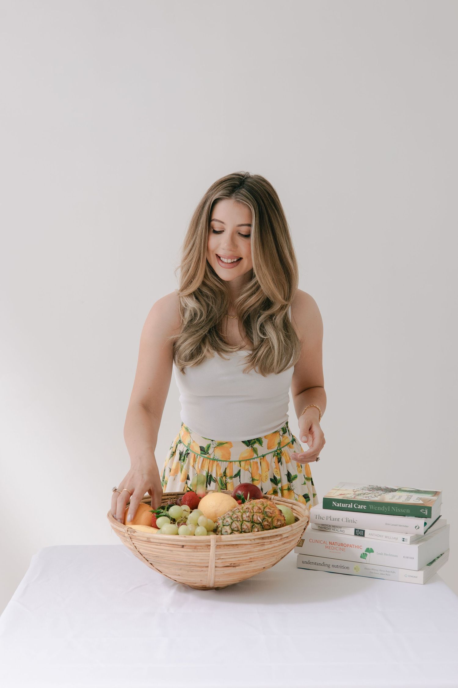 A person in a white tank top and floral skirt arranges fresh produce in a woven basket next to stacked cookbooks.