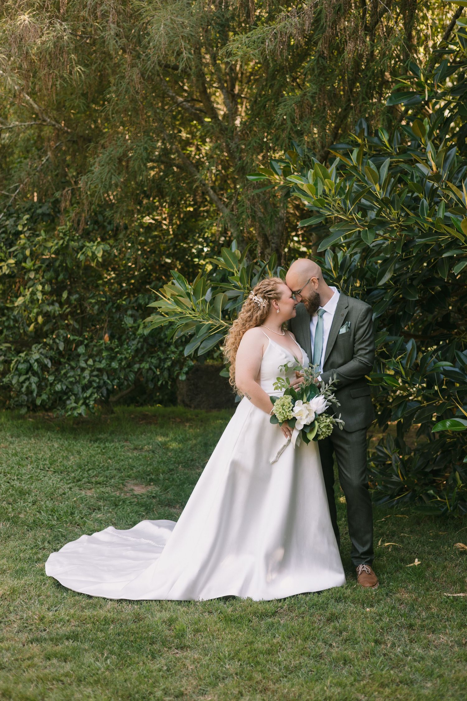 A romantic outdoor wedding portrait with a couple in formal attire standing next to lush greenery on a sunlit lawn.