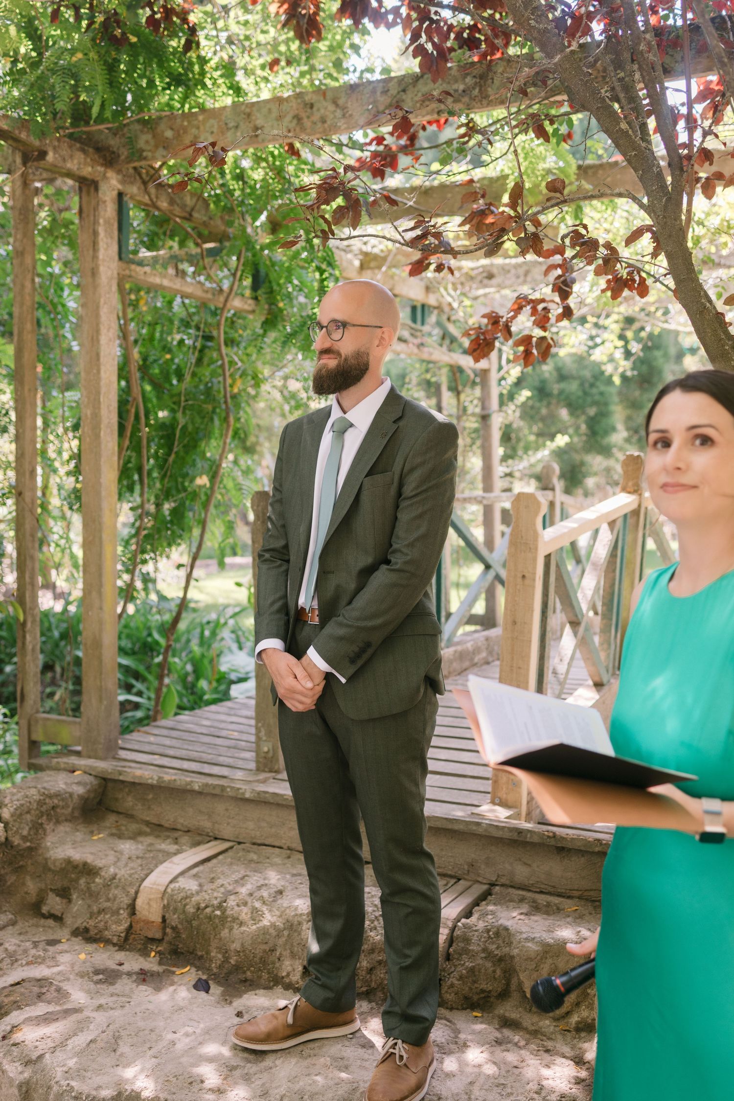 Wedding ceremony scene under a vine-covered pergola with a person in a suit and another in turquoise.