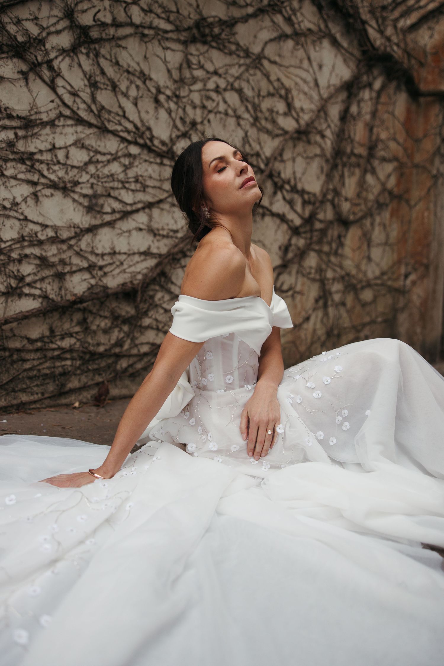A person in an elegant white wedding dress poses against a vine-covered wall with eyes closed in a dreamy moment.