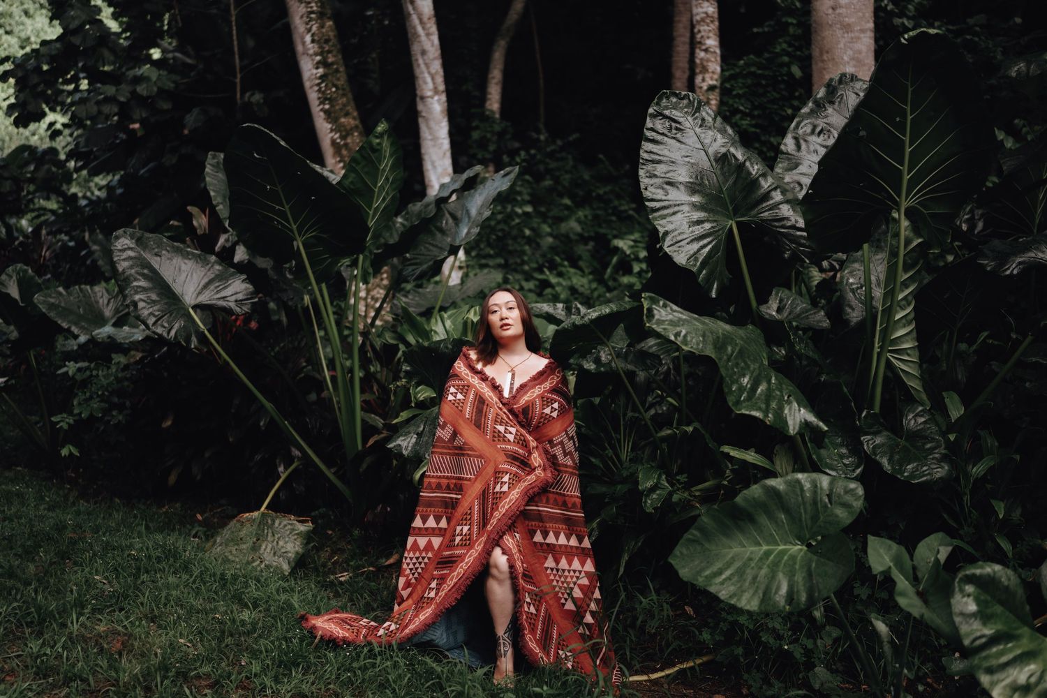A person sits wrapped in a patterned blanket surrounded by lush tropical plants and large elephant ear leaves in a garden.