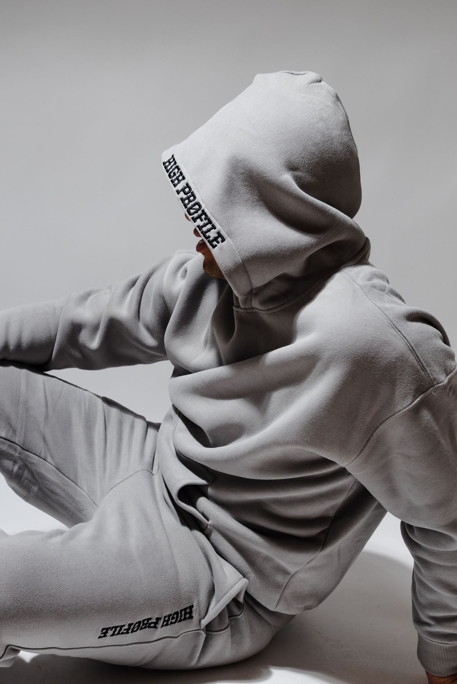 Athletic figure in light gray sweatsuit lounges against neutral background showing streetwear fashion.
